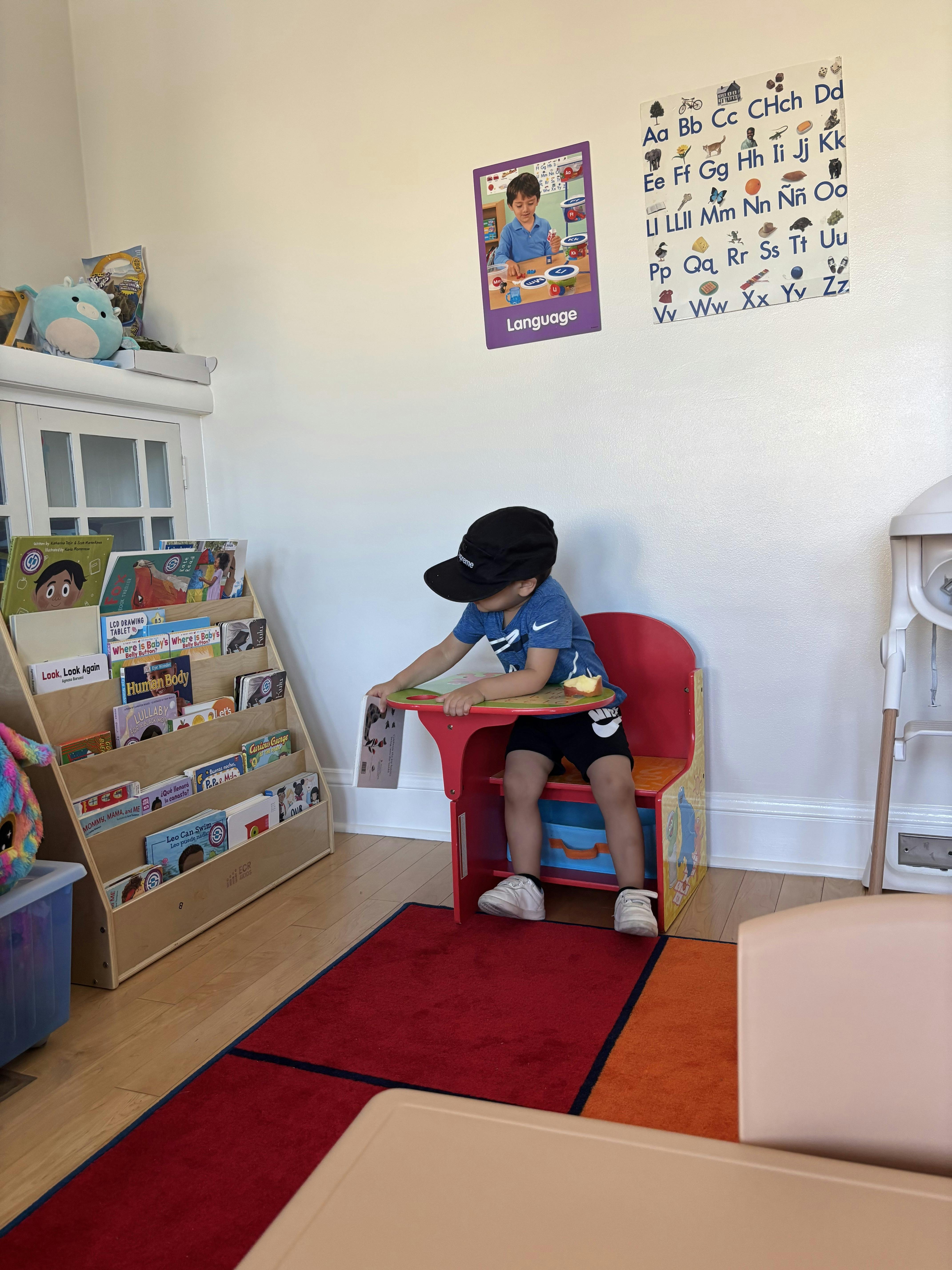 Child wearing a baseball cap sitting at a small red desk in a classroom