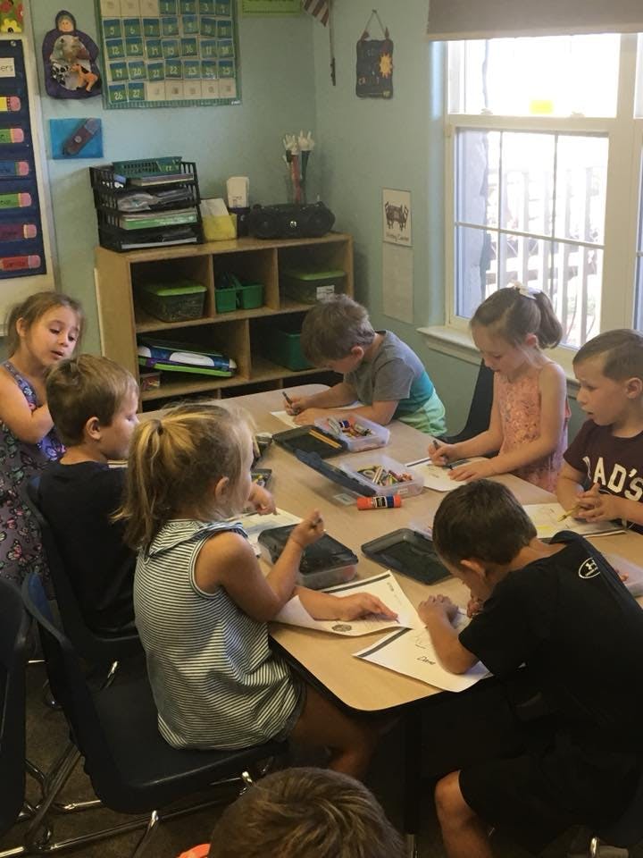 Children sitting at a table in a classroom, writing and drawing