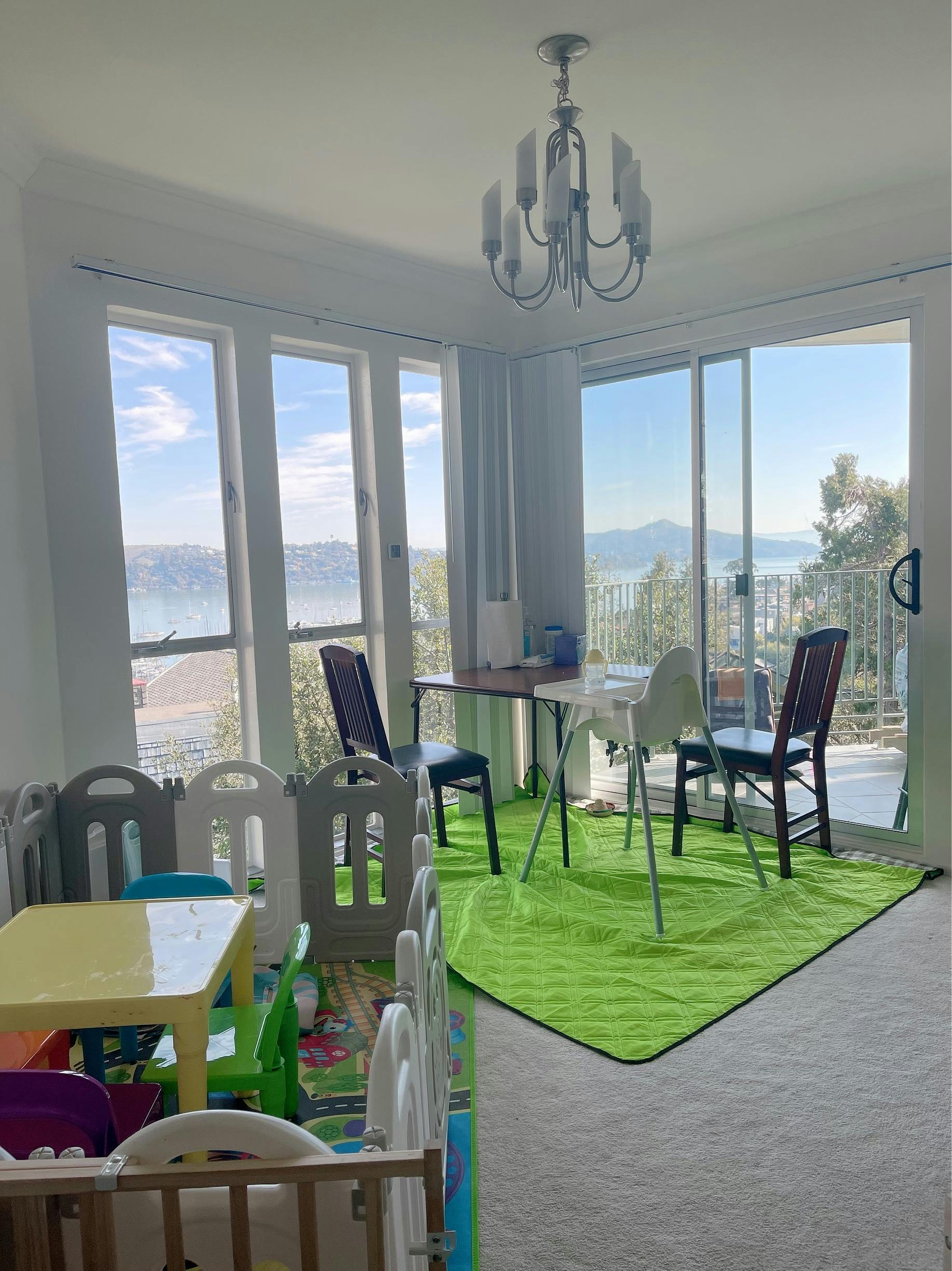 Room with large windows overlooking a body of water, with a playpen and dining table