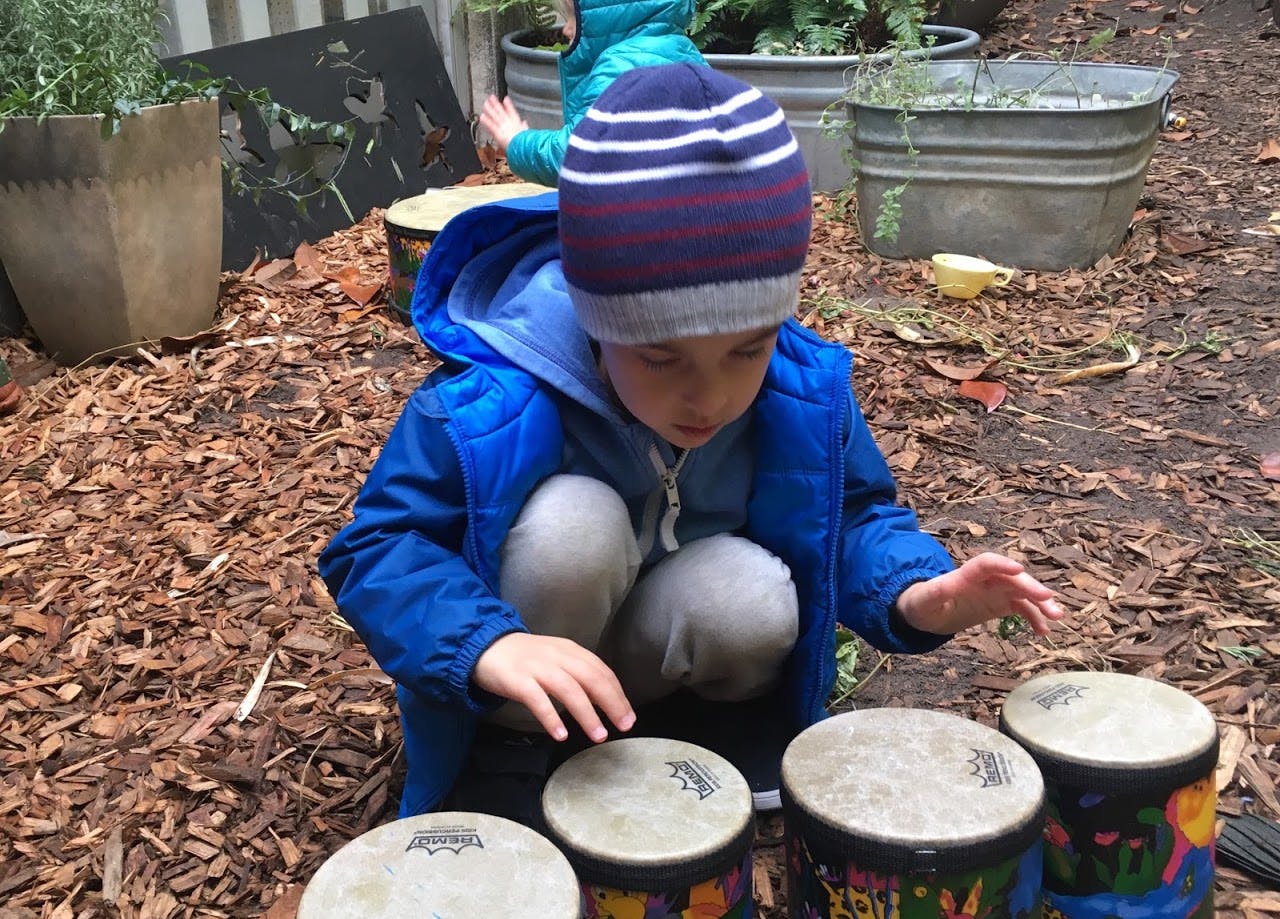Child in a blue jacket and striped hat playing bongo drums outdoors