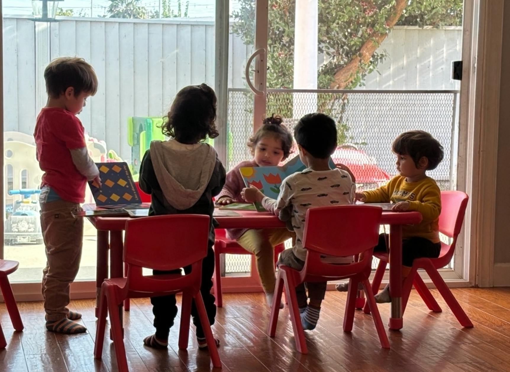 Children gathered around a small red table, one child standing with a laptop