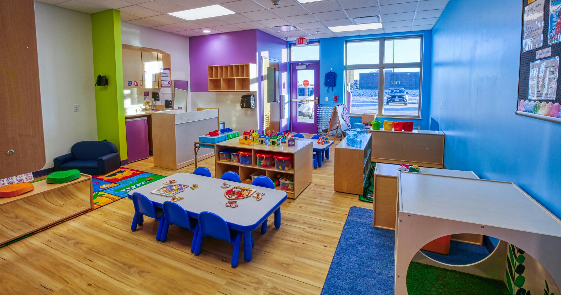 Colorful classroom with child-sized tables and chairs, shelves with toys, and large windows