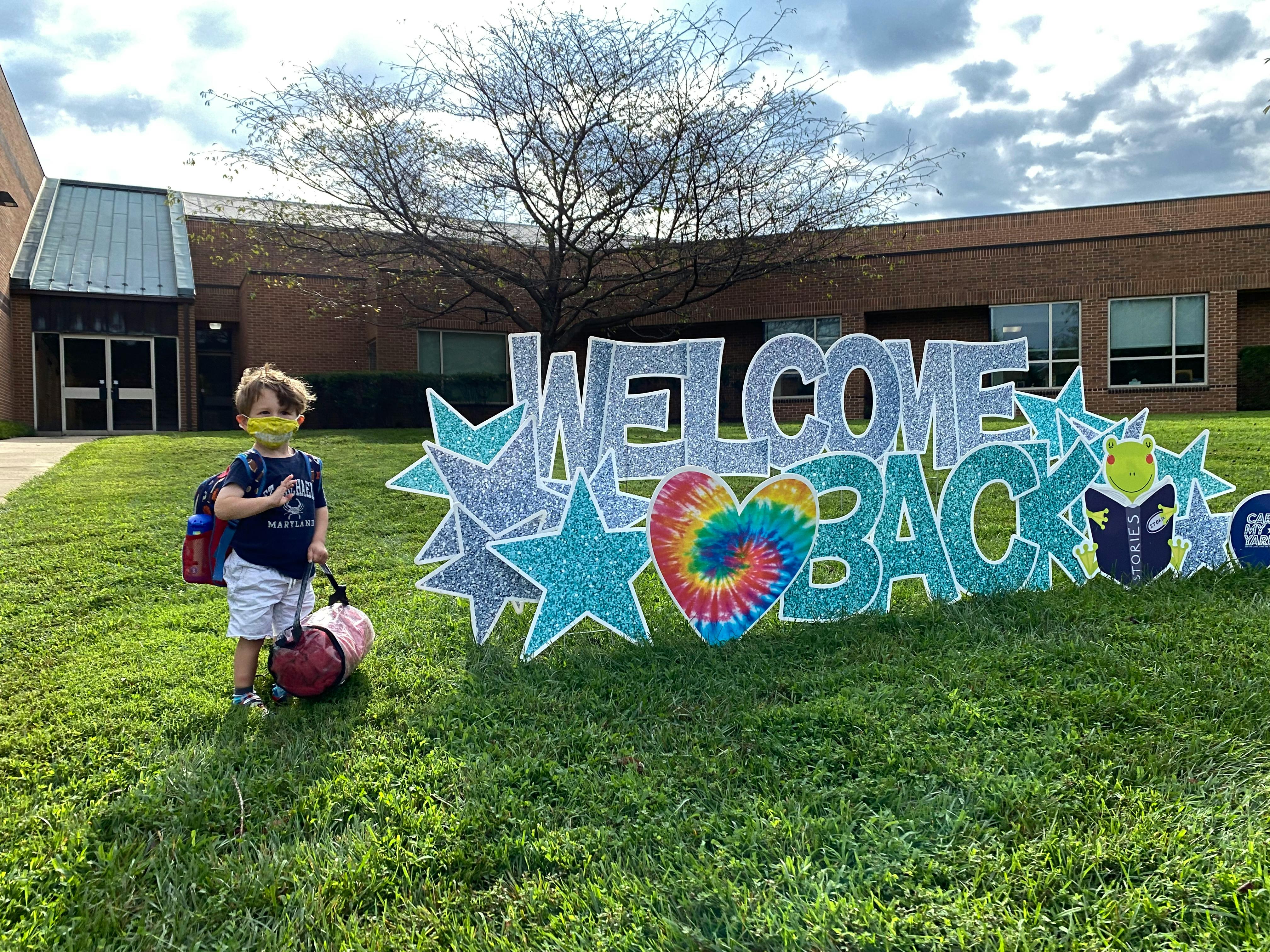 Child with backpack and bag standing next to a "Welcome Back" sign
