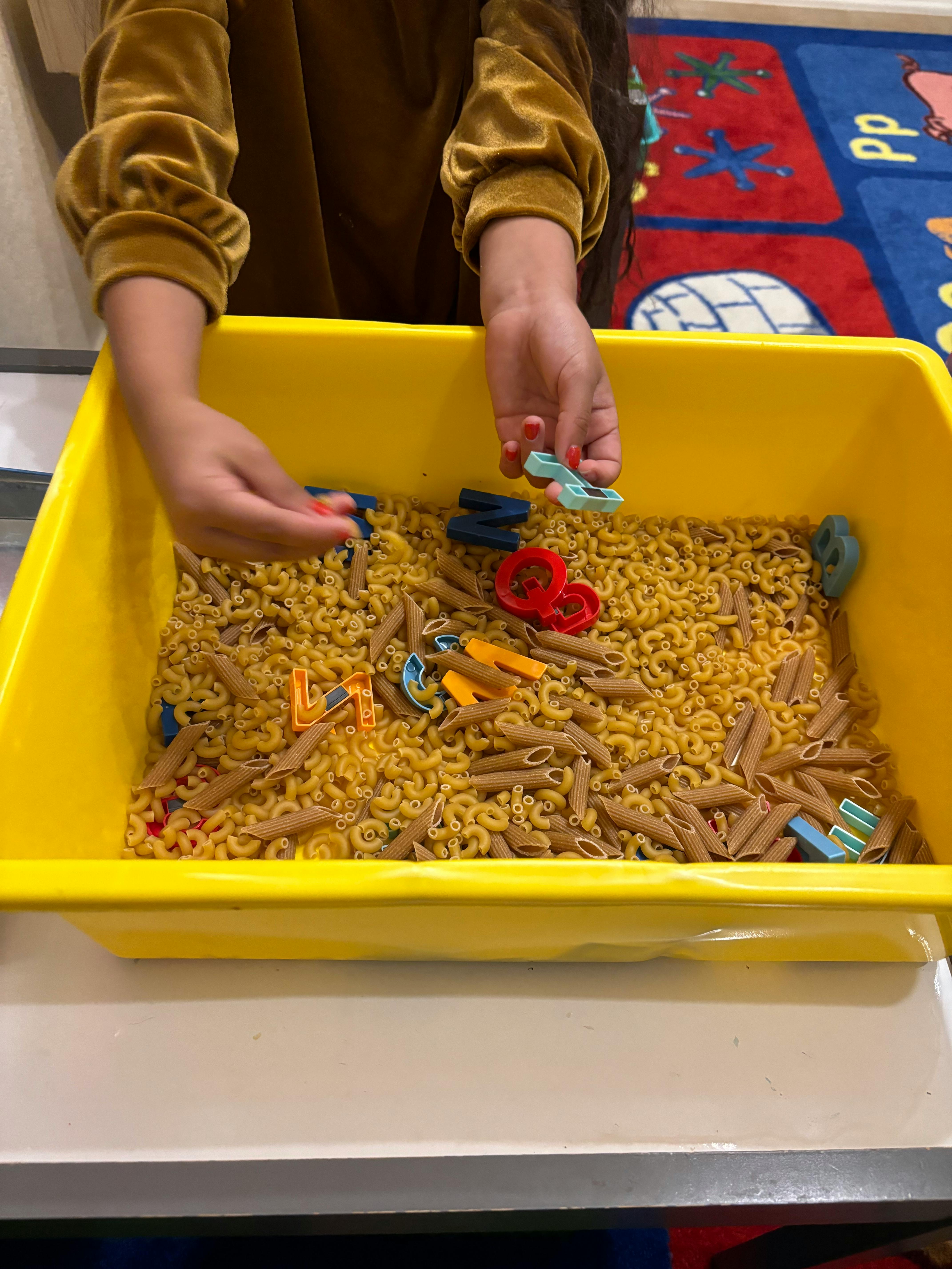 Child's hands playing with pasta and letter shapes in a yellow bin