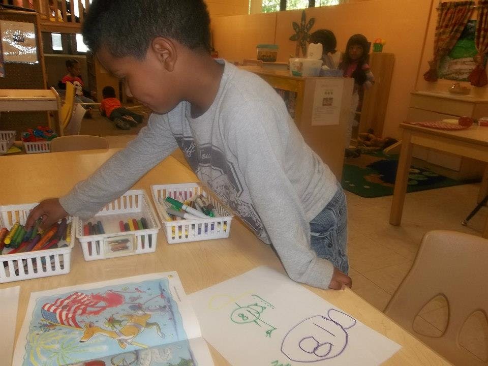 Child reaching into a basket of art supplies on a table with drawings on paper