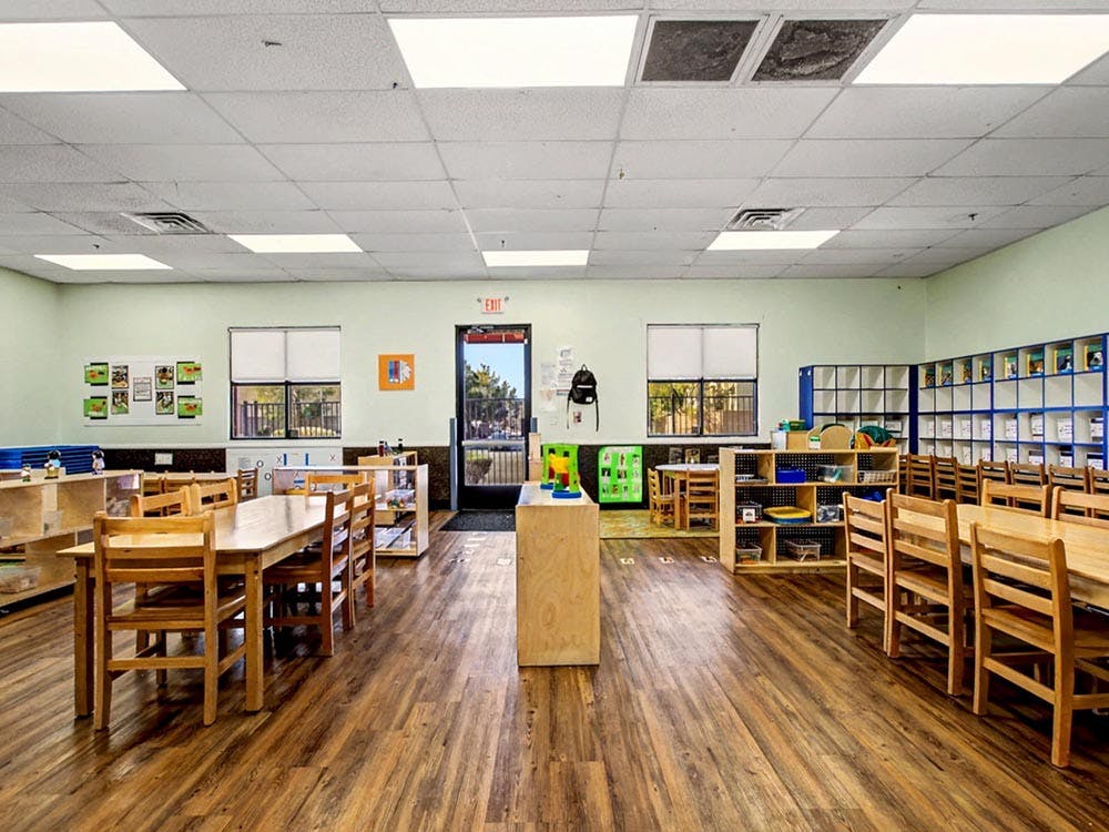 Empty classroom with small wooden tables and chairs, shelves, and a door with an exit sign