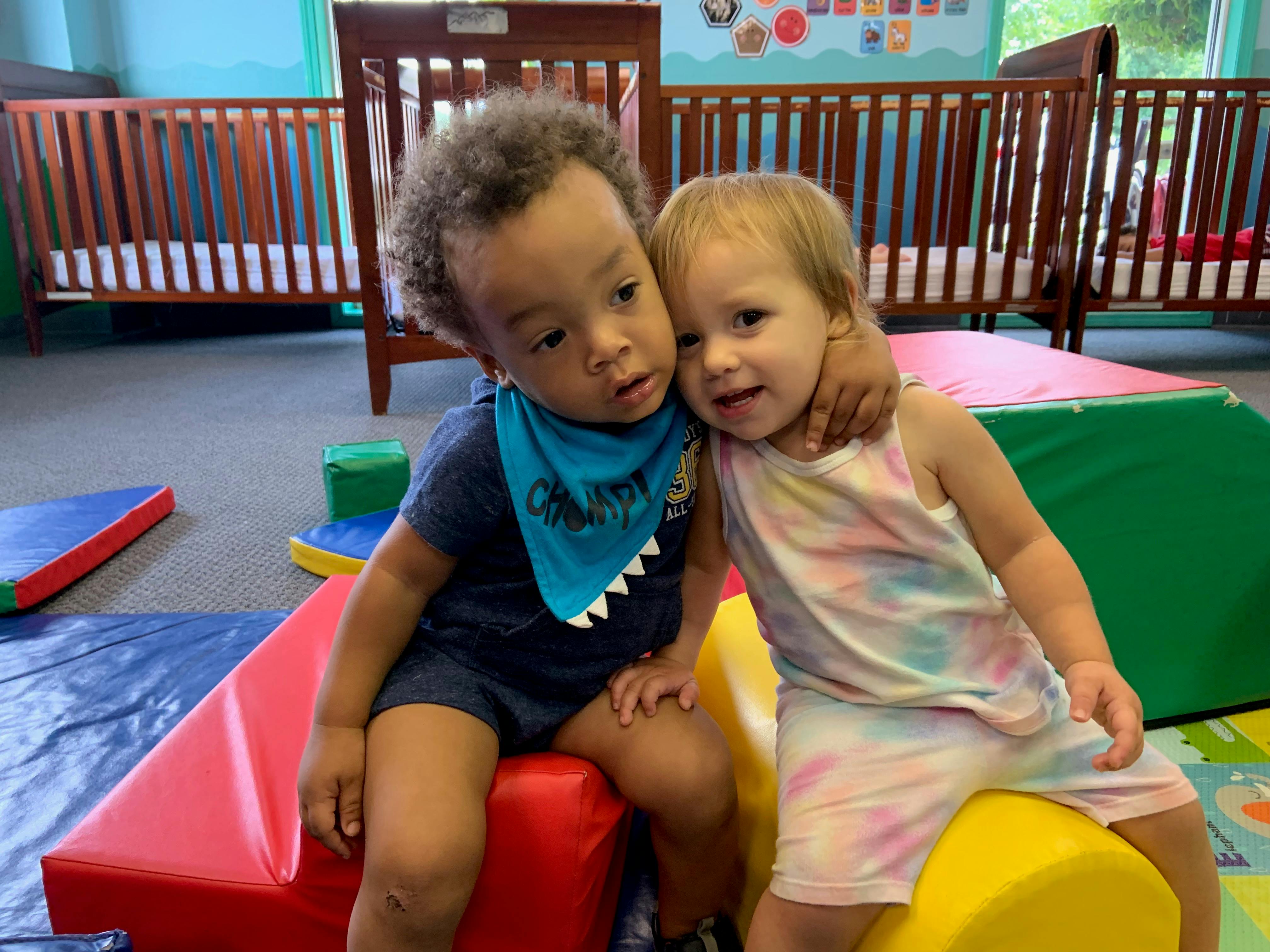 Two young children hugging each other while sitting on play mats in a room with cribs