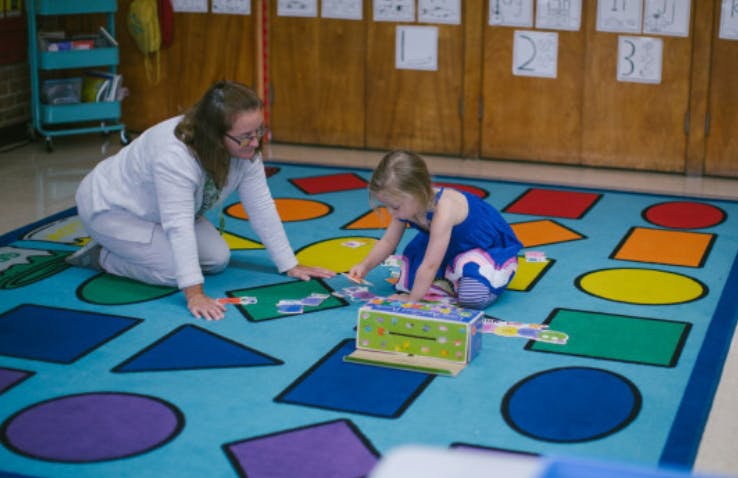 Person and child kneeling on a rug with shapes, playing with a toy