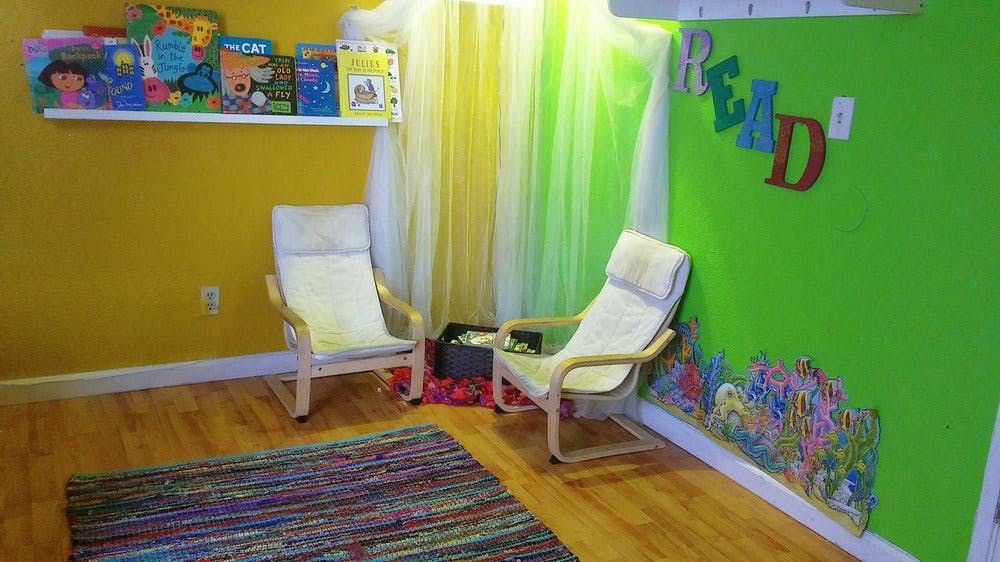 Two chairs in a reading nook with books on a shelf and a rug on the wall