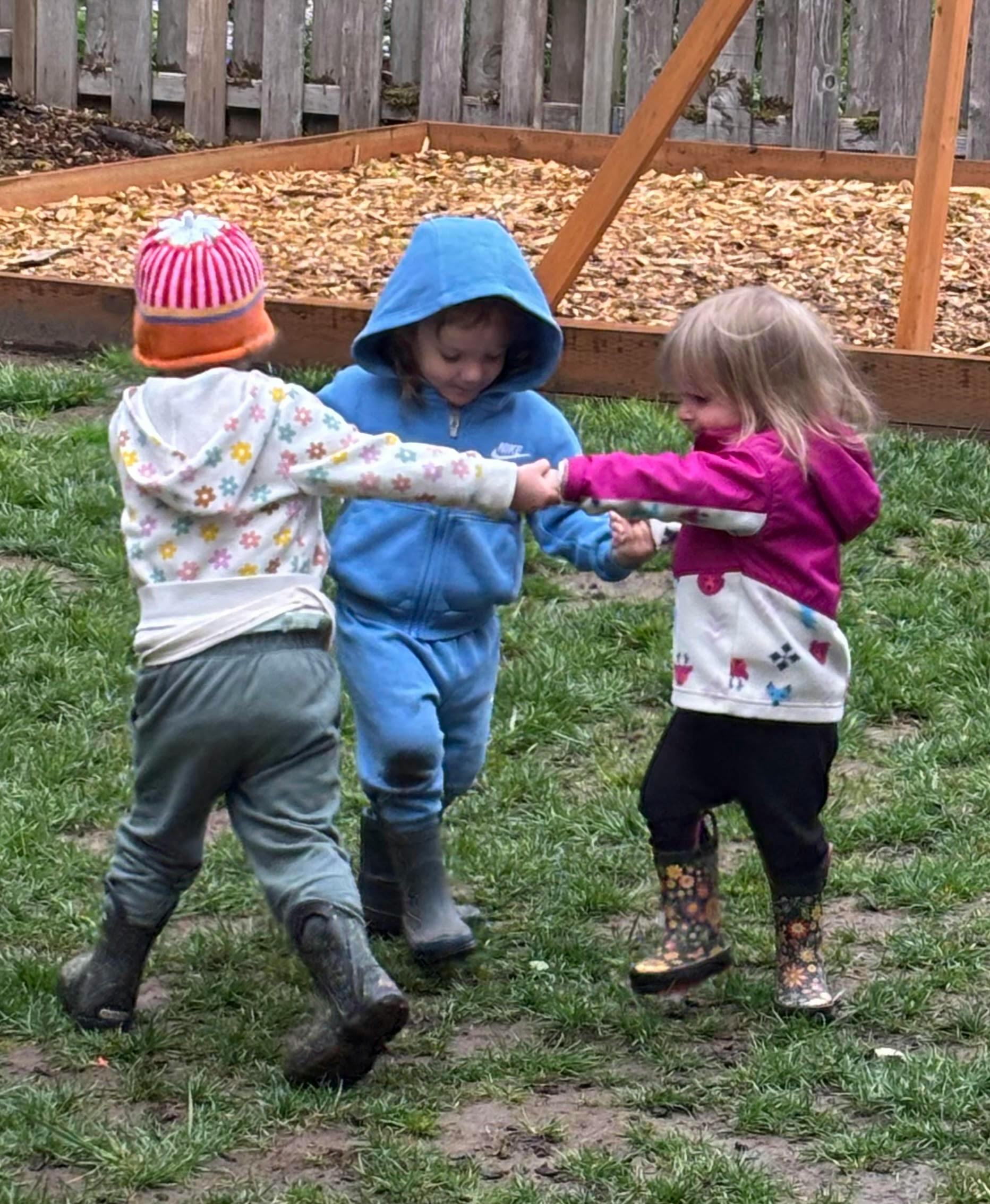 Three children holding hands in a circle on grass, wearing rain boots