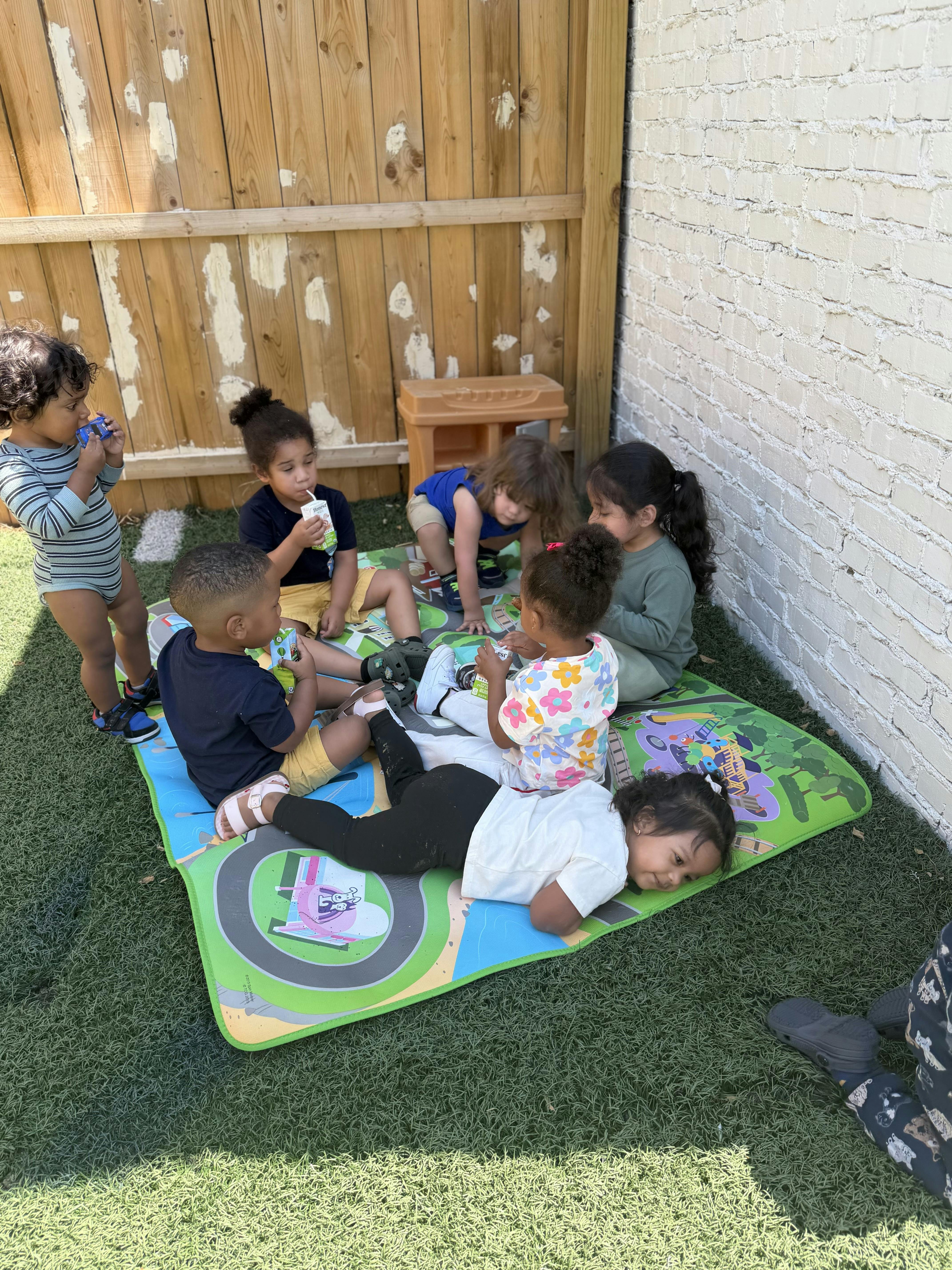Group of children on a play mat outdoors, some drinking from juice boxes