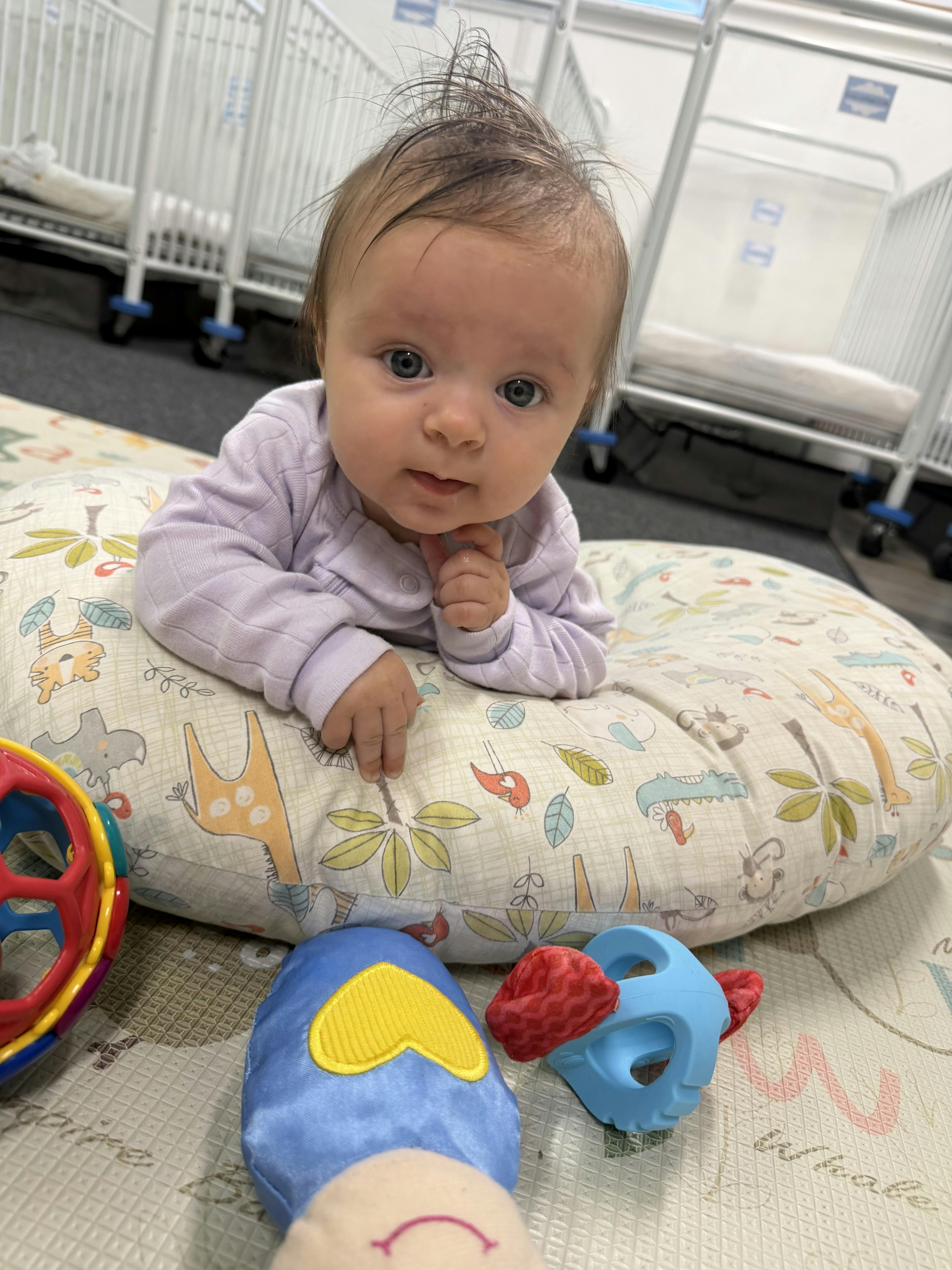 Baby lying on stomach on a patterned support pillow with toys nearby