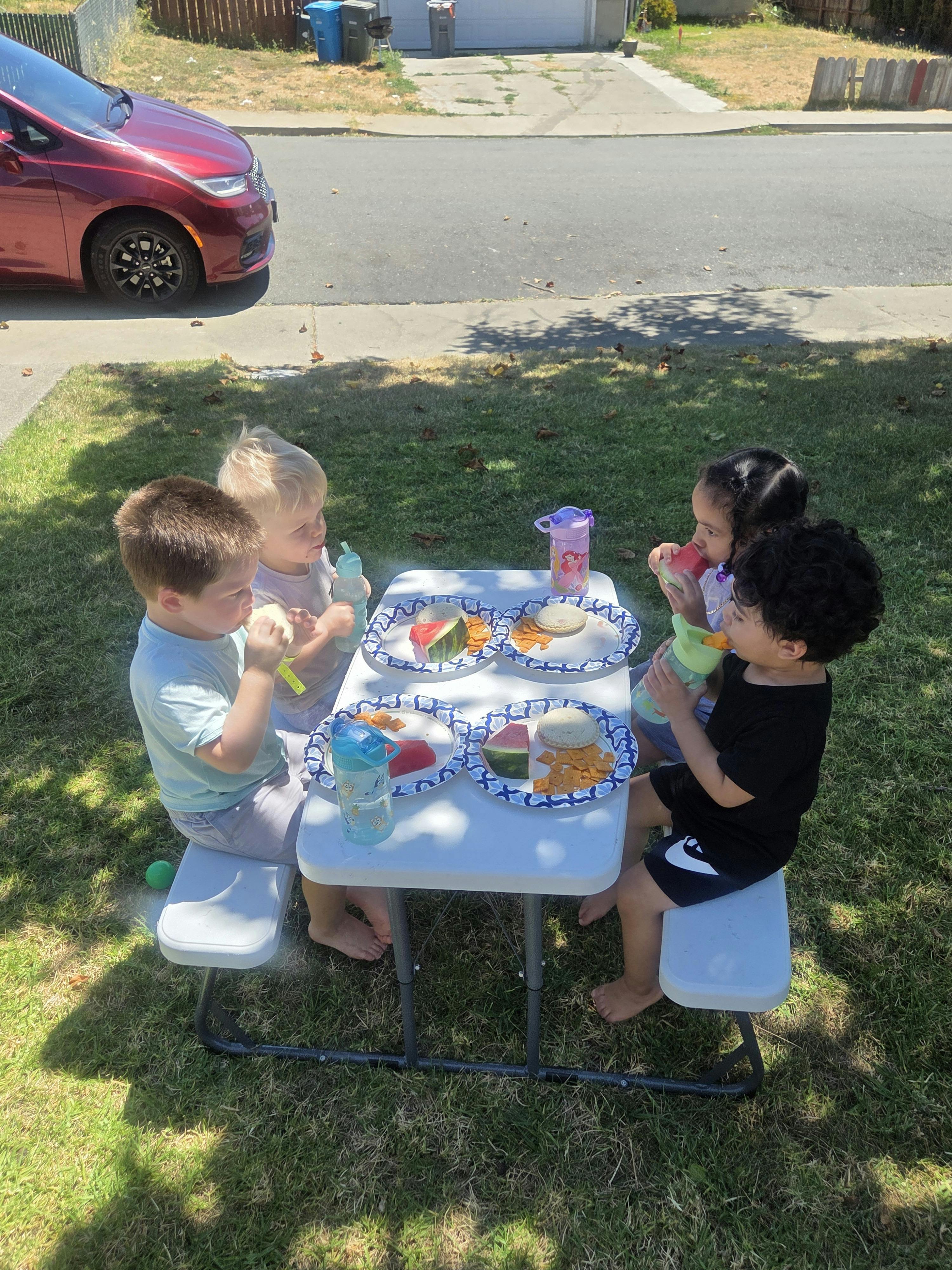 Four children eating lunch at a small plastic table on a grassy lawn