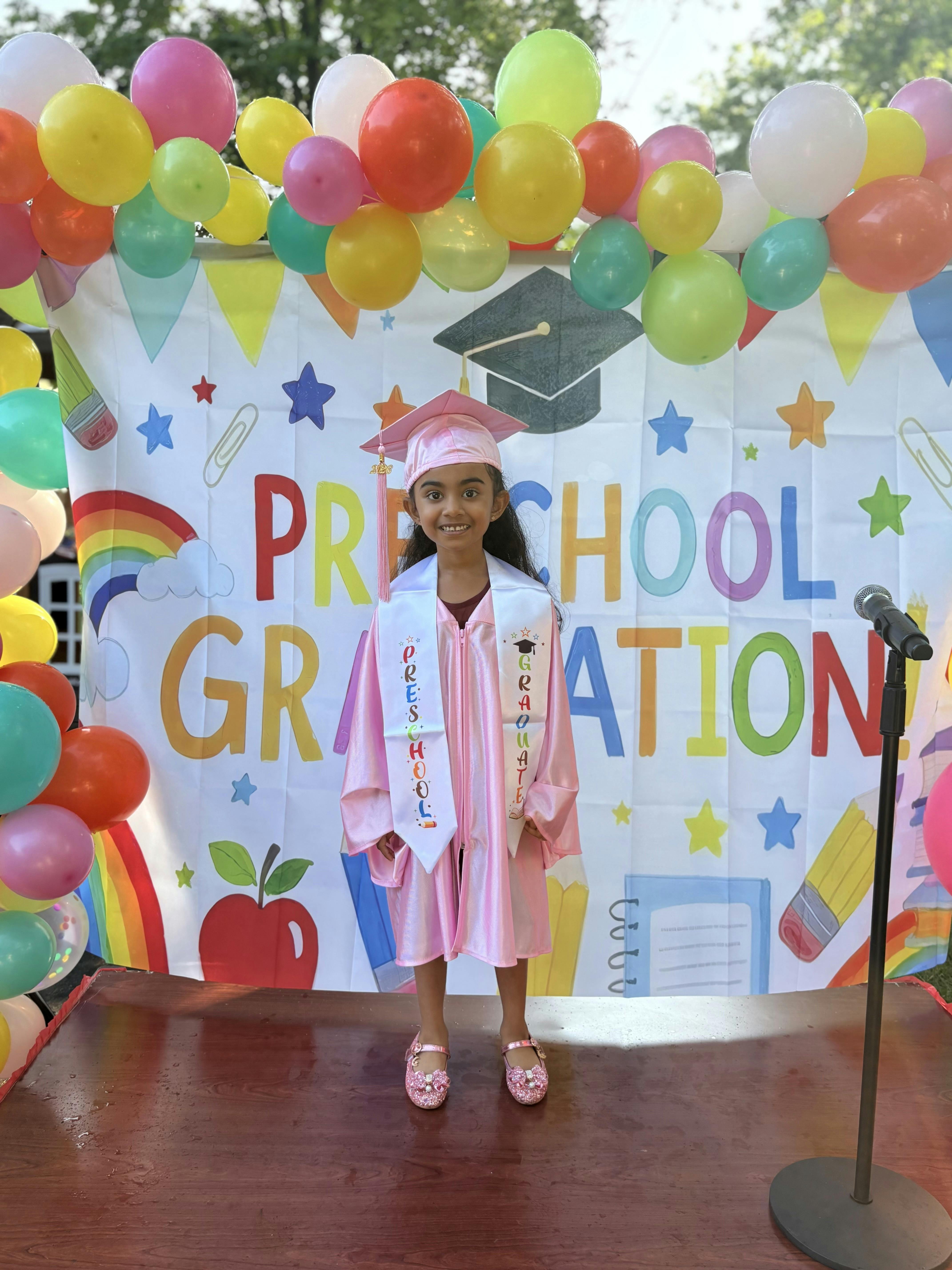 Child wearing pink preschool graduation cap and gown in front of decorated banner
