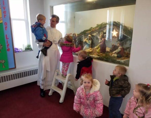 Adult holding a child, with other children looking at a nativity scene