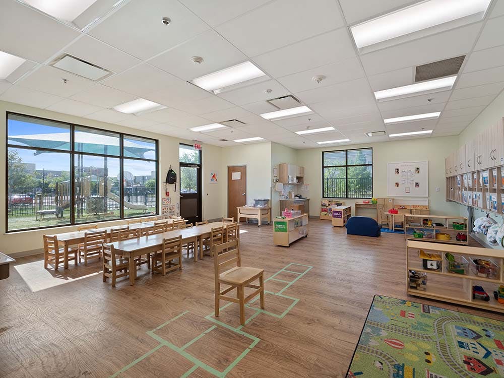 Empty classroom with small wooden tables and chairs, toys, and large windows