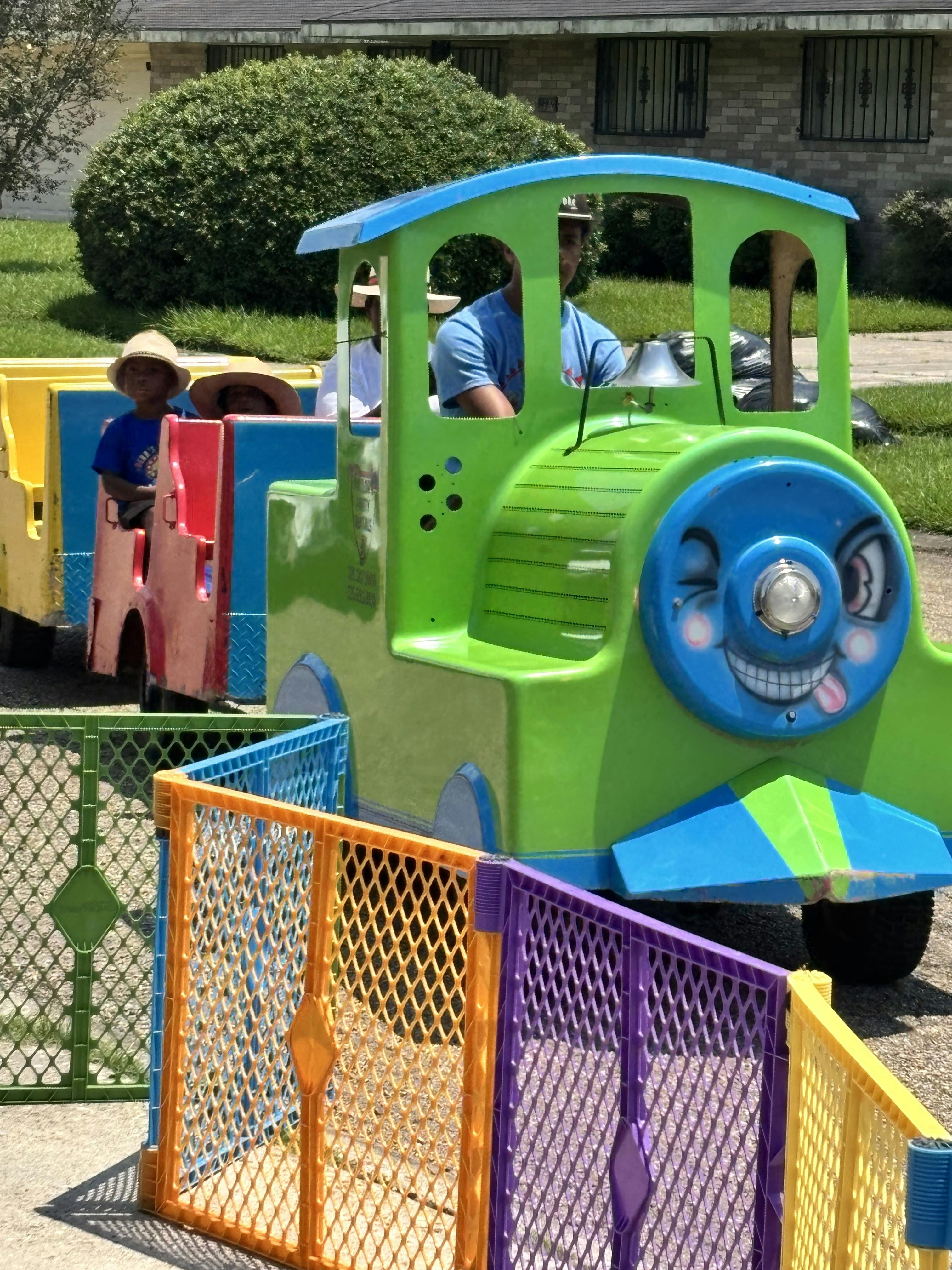 People riding in a green and blue toy train with colorful fences