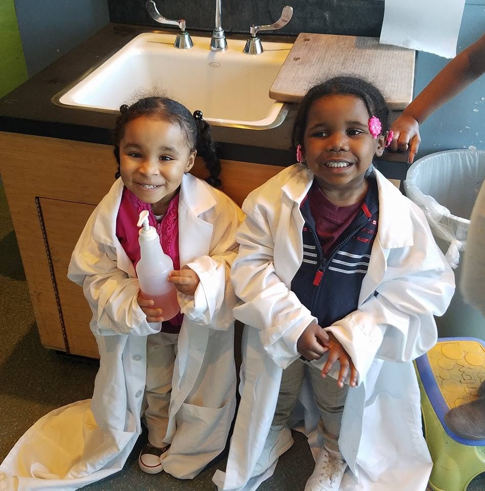 Two children wearing lab coats standing in front of a sink