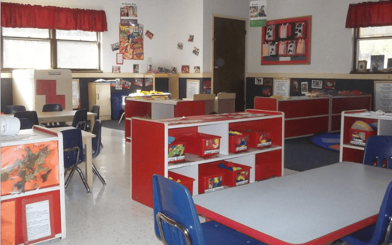 Classroom with small tables and chairs, shelves with red bins, and bulletin boards