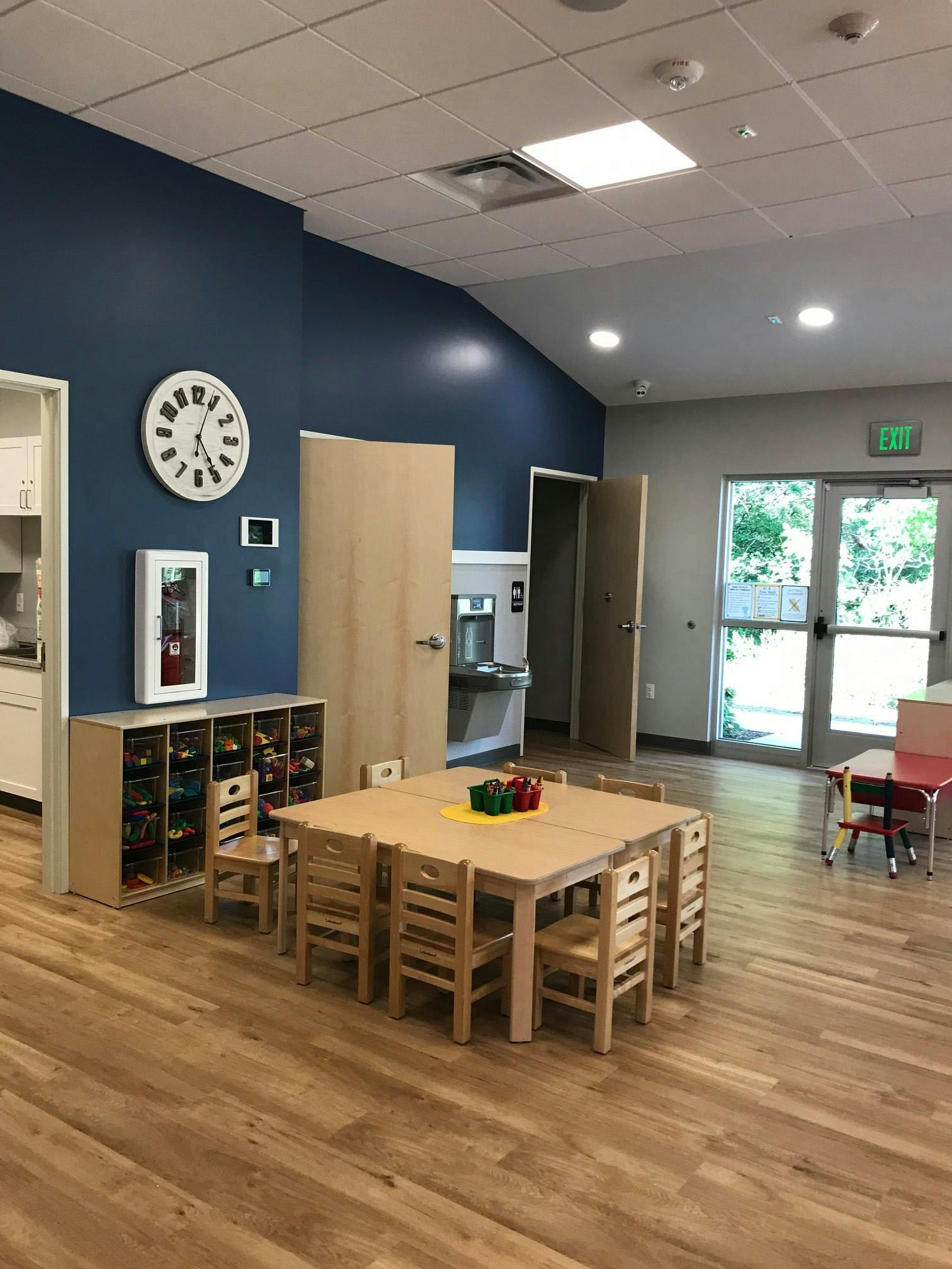 Classroom with small wooden tables and chairs, blue accent wall, and large windows