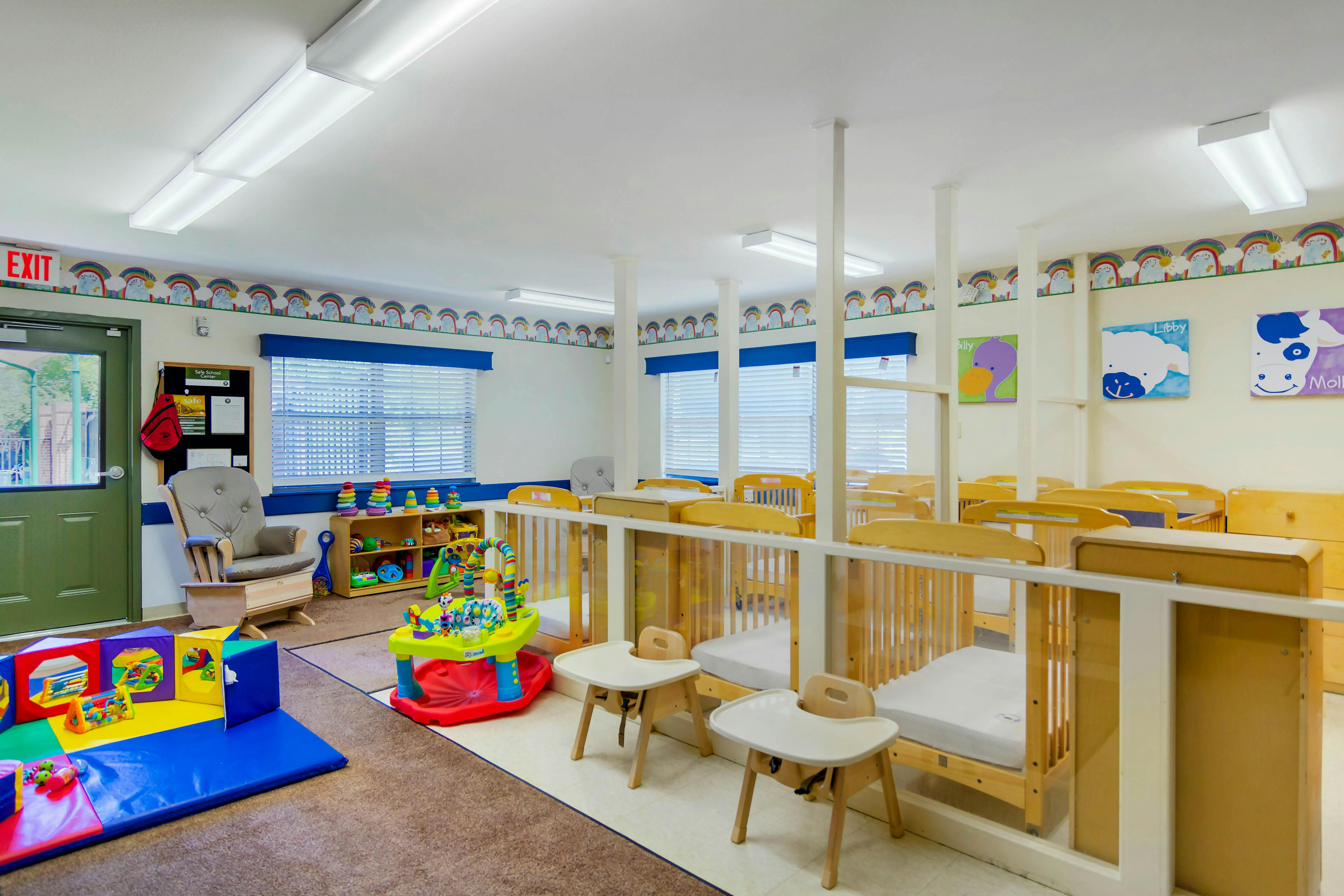 A colorful childcare room with cribs, playmats, toys, and a rocking chair