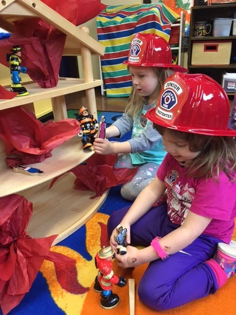 Two children in red firefighter hats playing with toys on the floor
