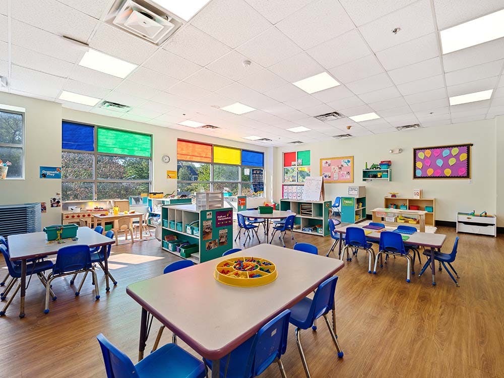 Empty classroom with small tables and chairs, colorful blinds on large windows