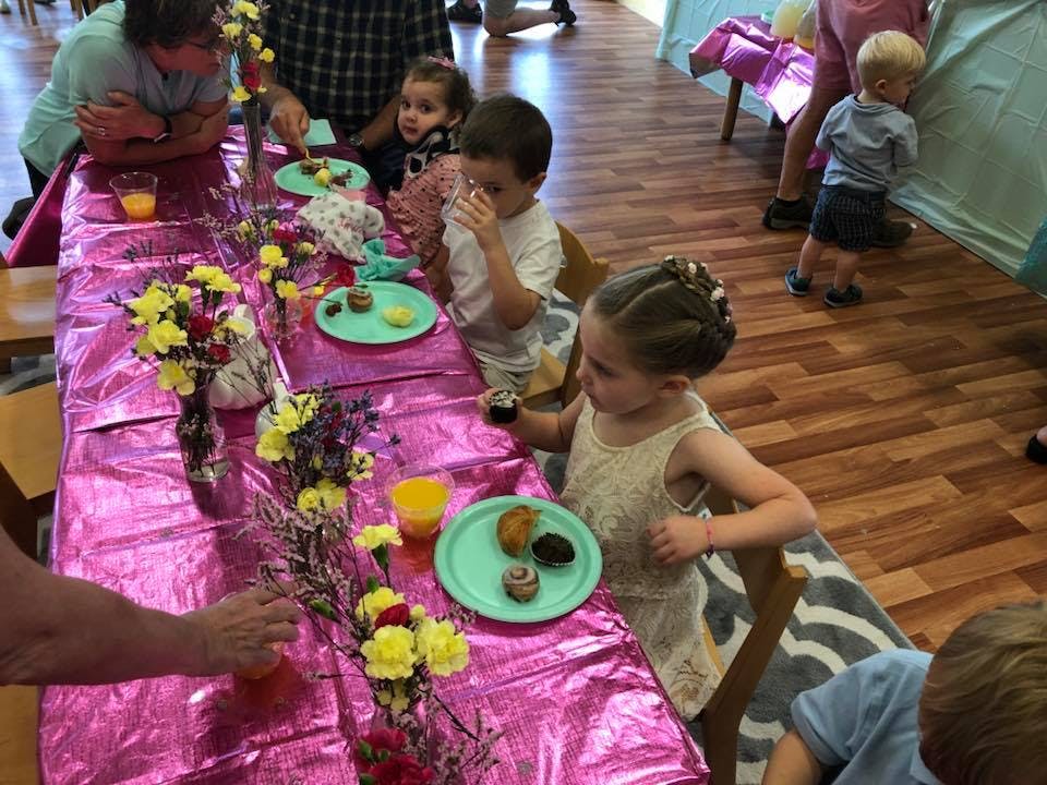 Children and adults seated at a long table with flowers and plates of food