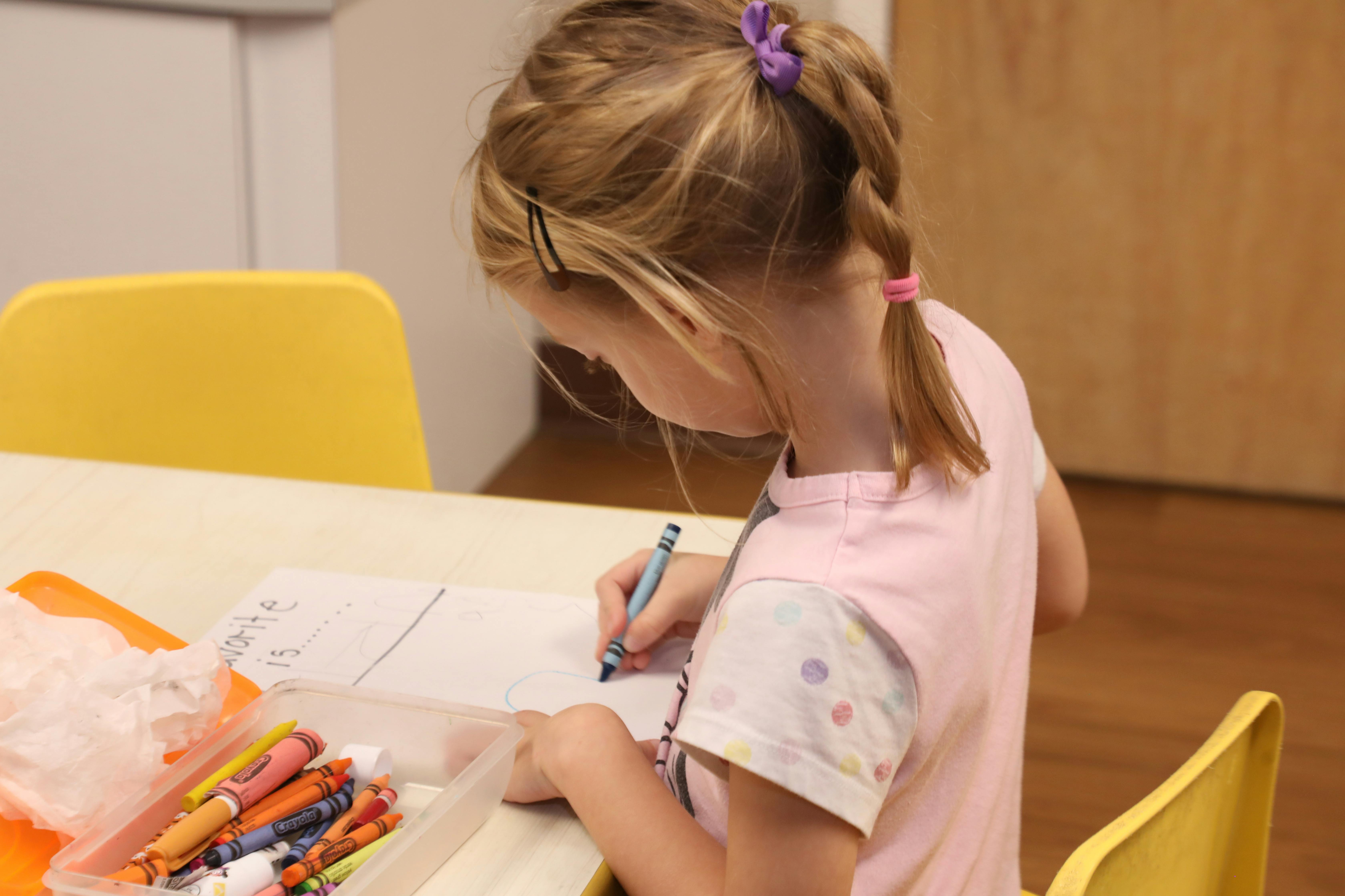 Child writing on paper at a table with crayons