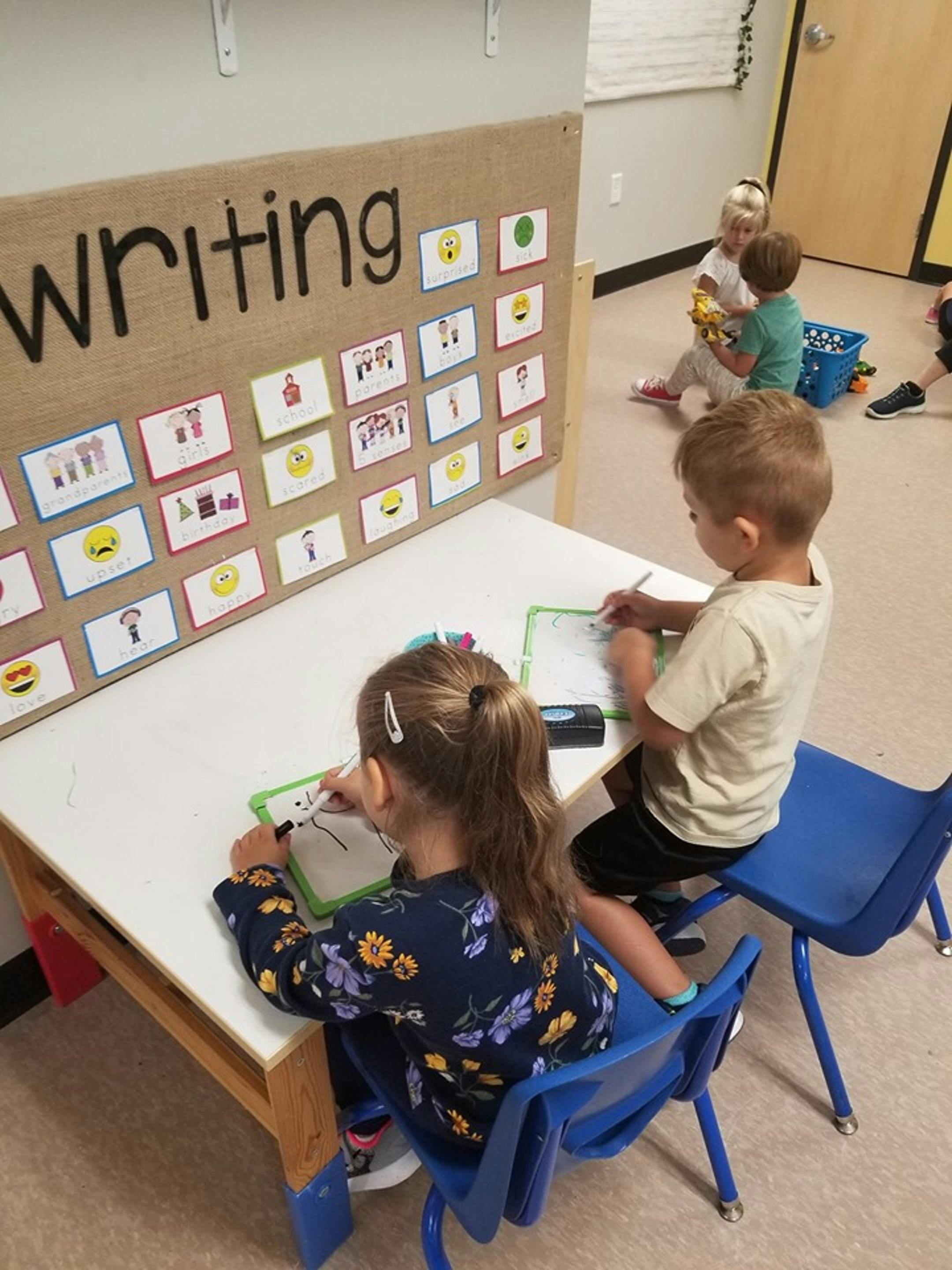 Two children writing at a table with a "Writing" sign in the background