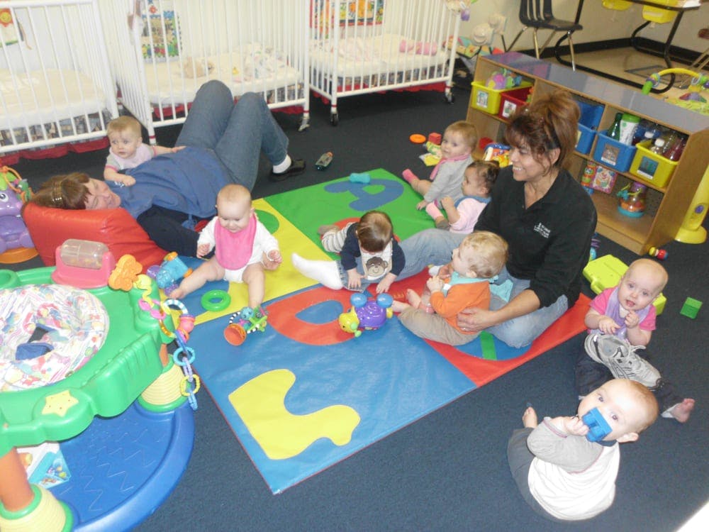 Two people with multiple children playing on mats in a room with cribs in the background