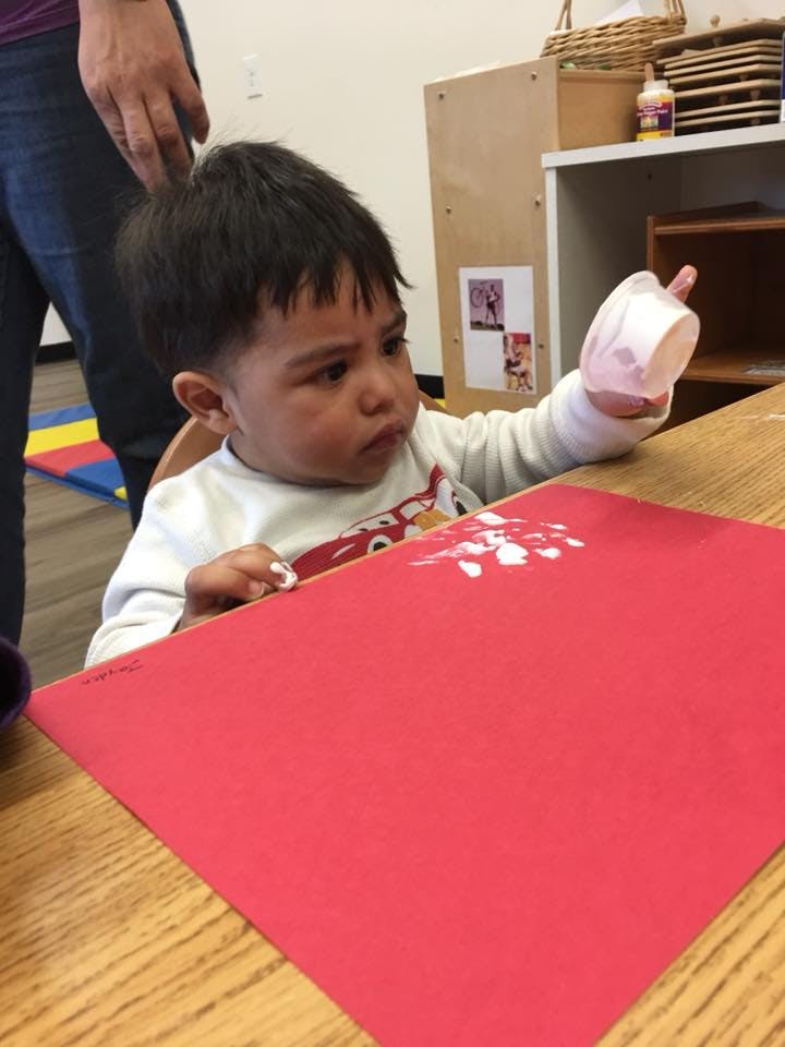 Child holding a cup over a red paper with white paint on it