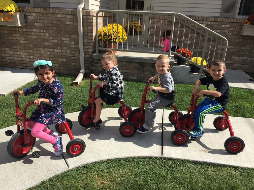 Four children riding red tricycles on a sidewalk