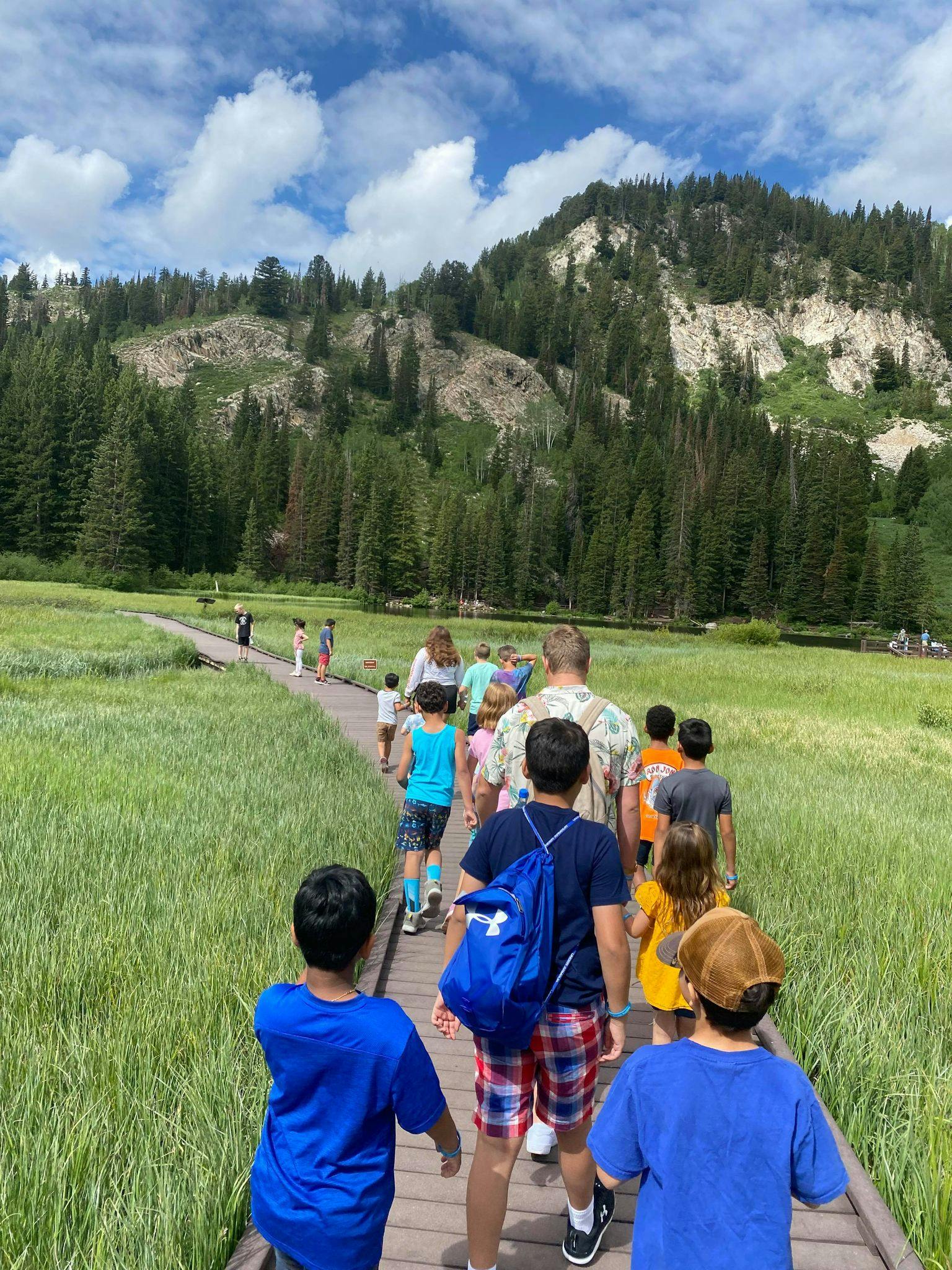 People walking on a boardwalk through a grassy field with mountains in the background