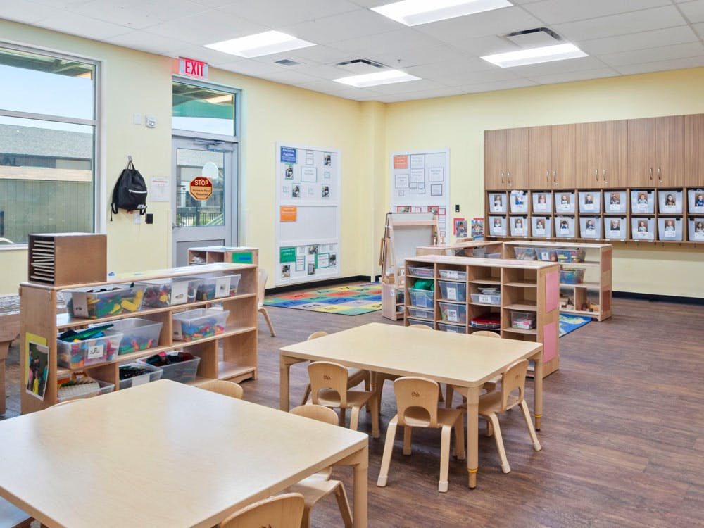 An empty classroom with small tables and chairs, shelves with toys, and cabinets