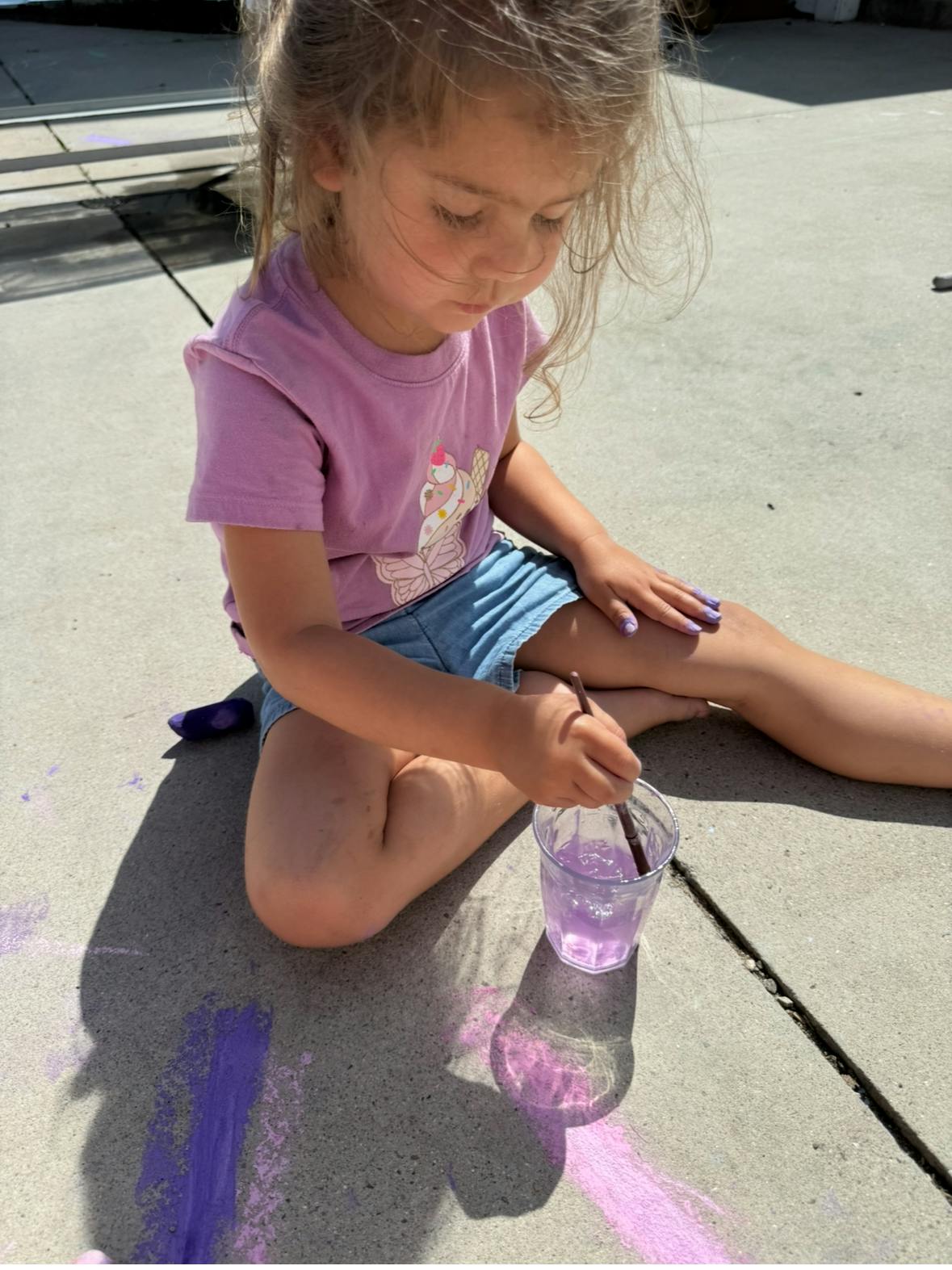 Child sitting on pavement, painting with a brush and purple liquid