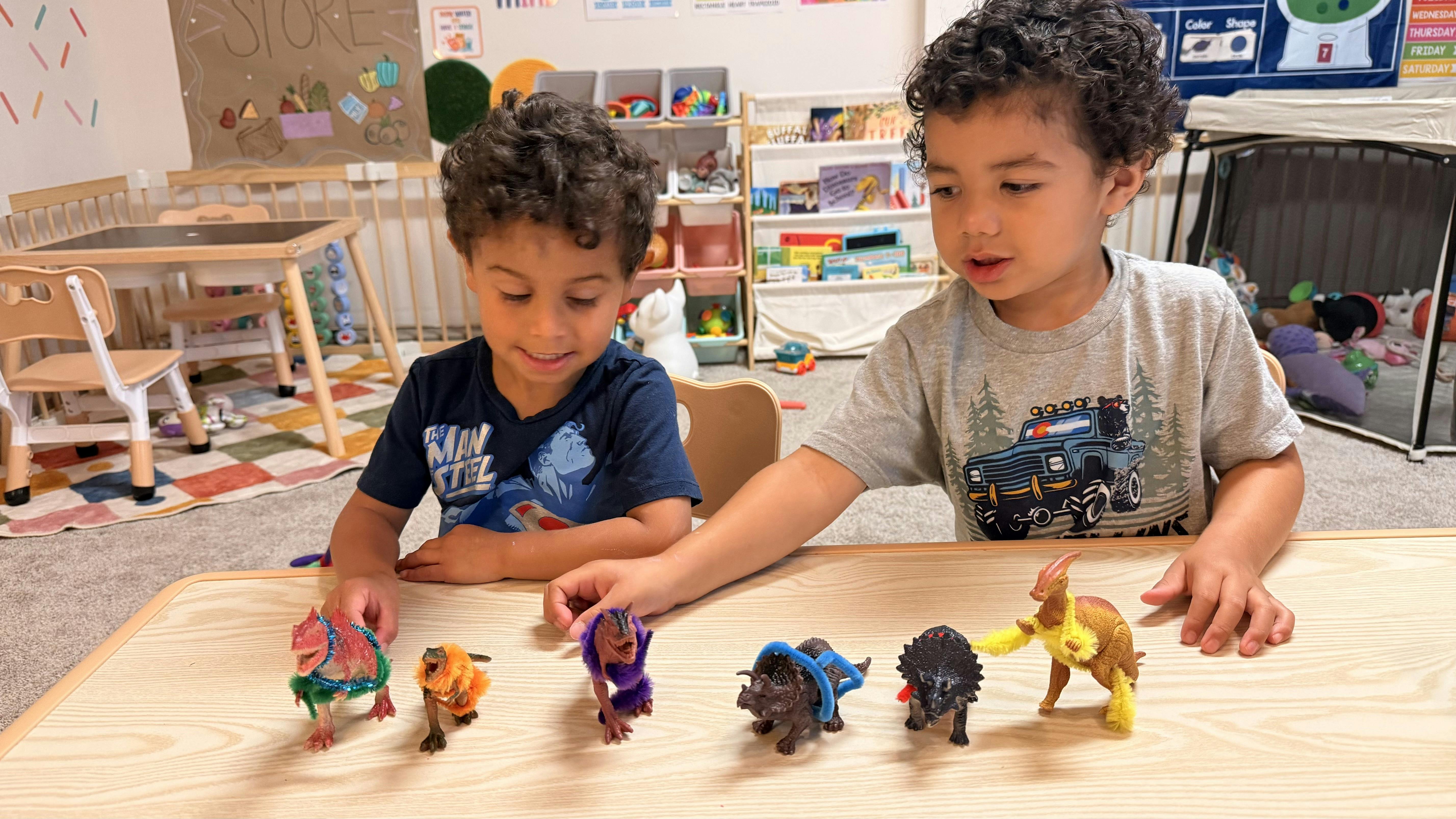 Two children playing with dinosaur figures at a table in a classroom