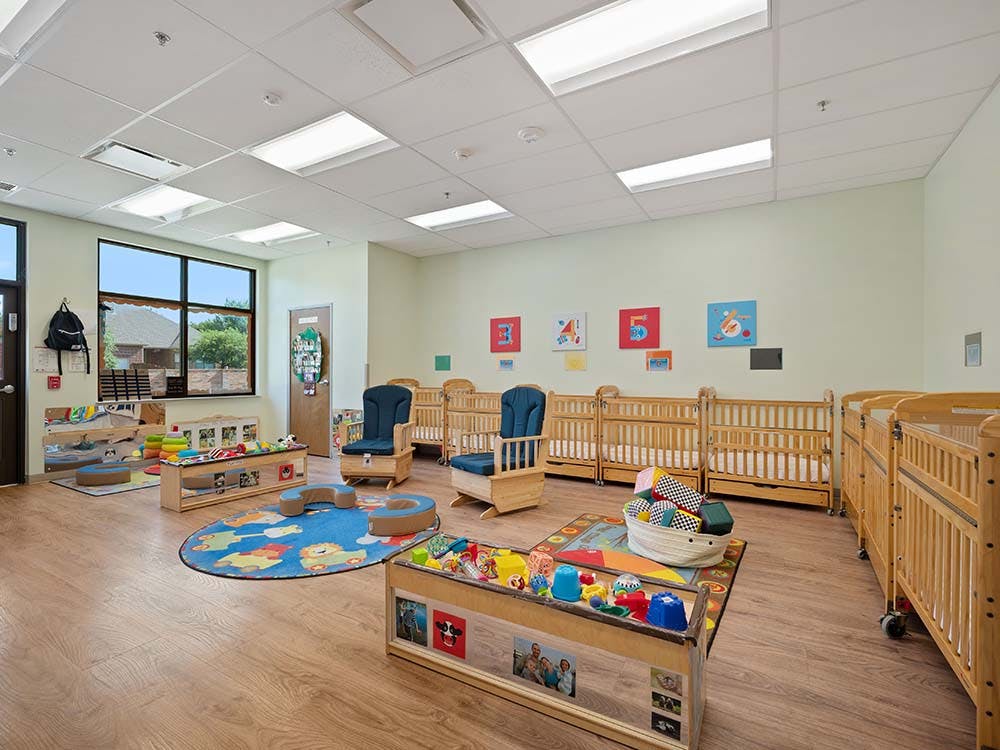 Indoor infant or toddler classroom with cribs, play rug, and activity center