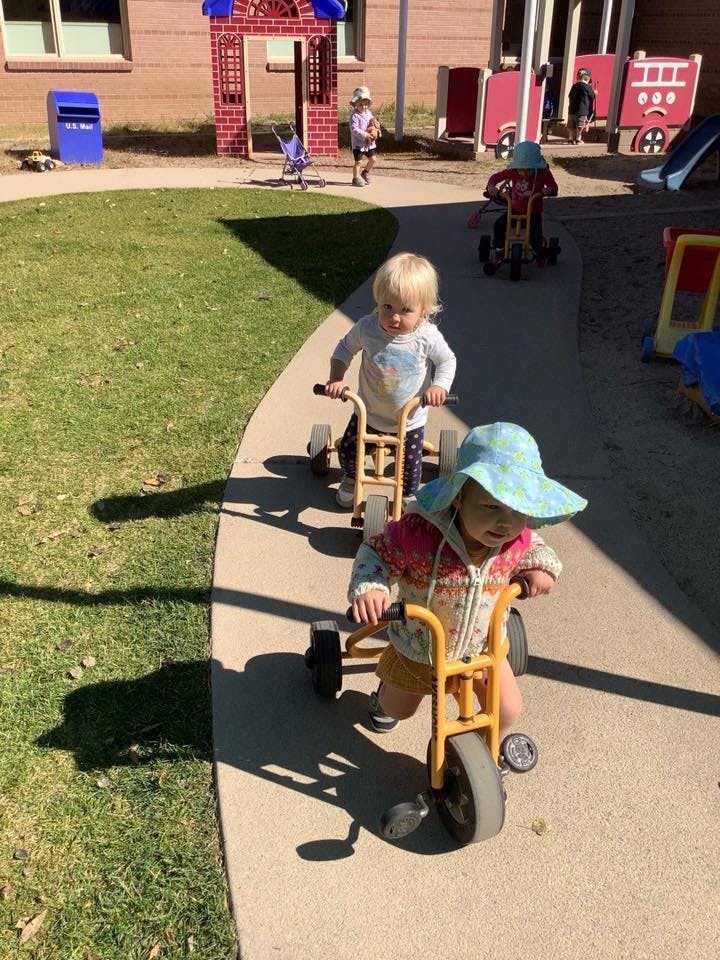Two children riding tricycles on a paved path outdoors