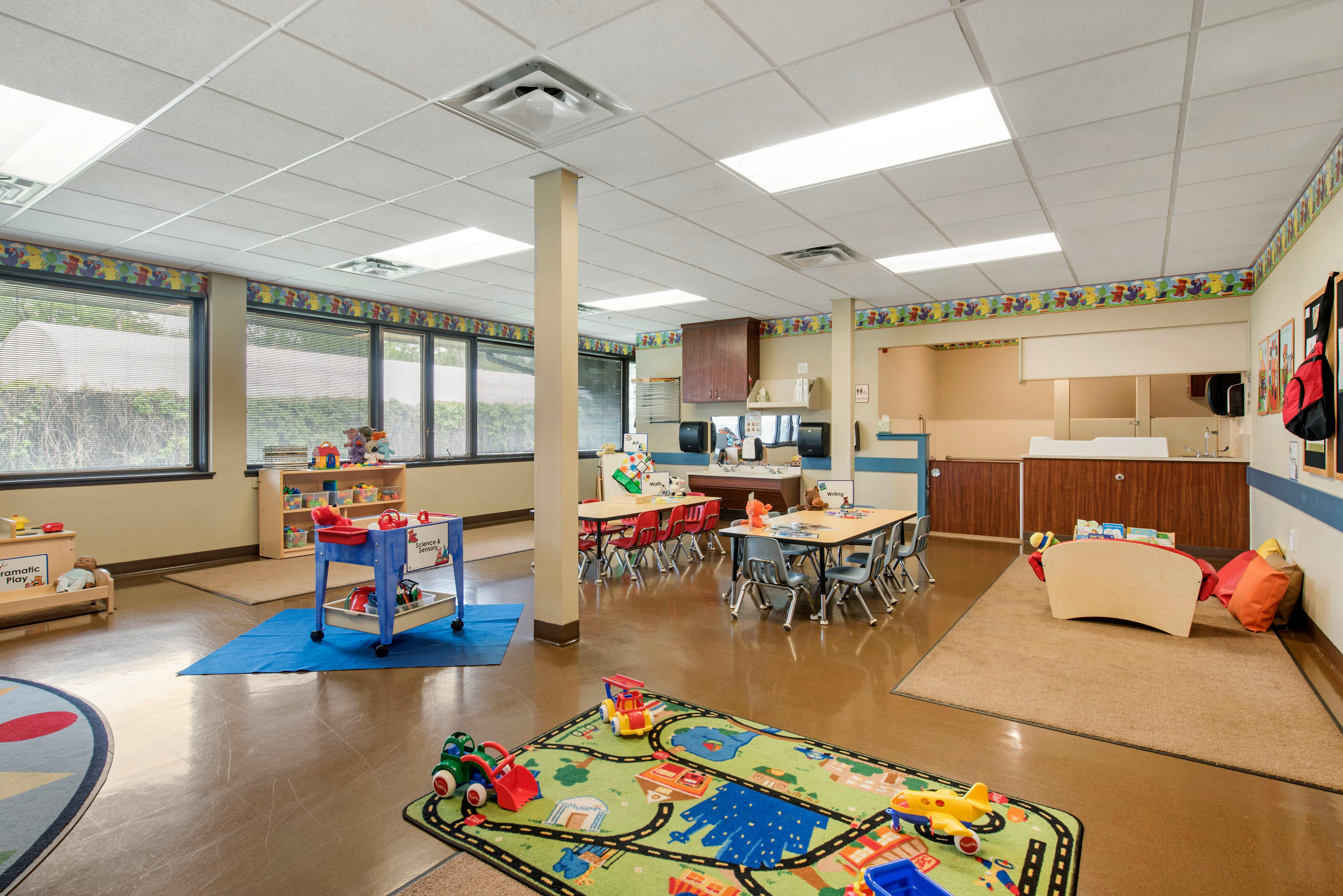 Classroom with small tables and chairs, play mats, toys, and windows