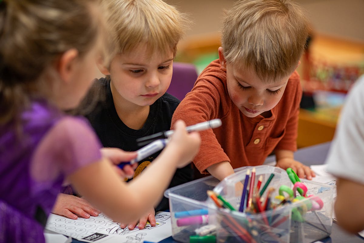 Three children drawing and selecting markers from a container