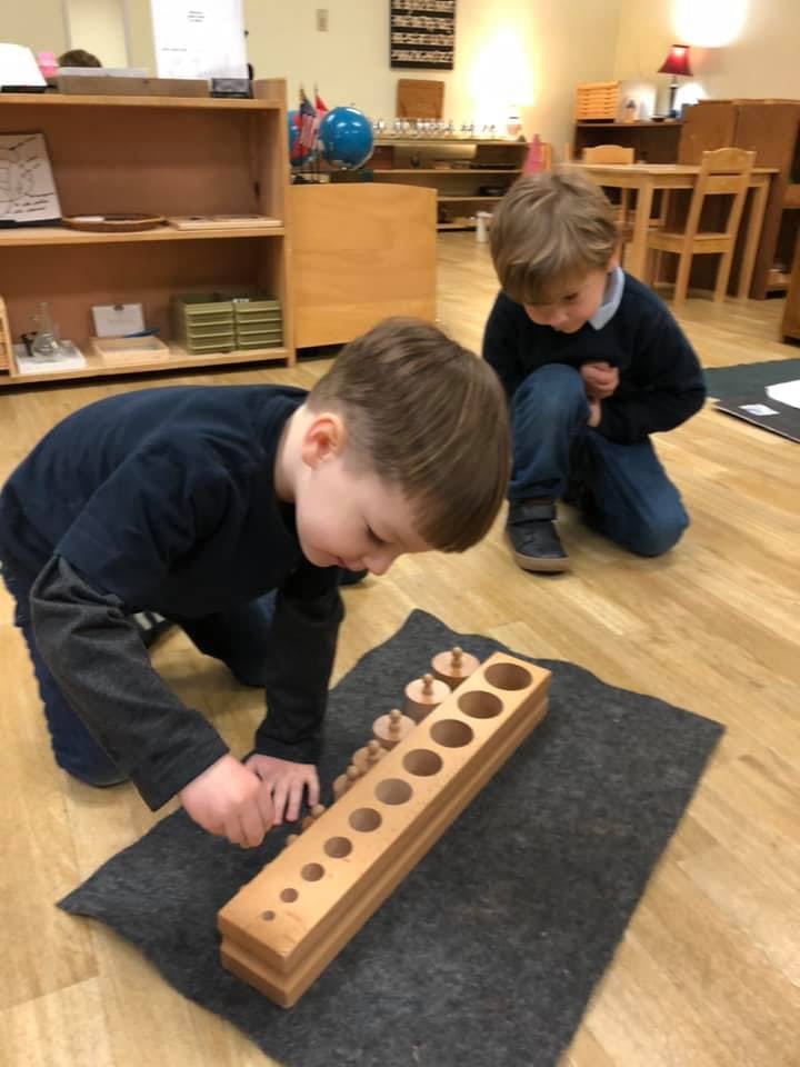 Two children kneeling on the floor, one playing with a wooden cylinder block set