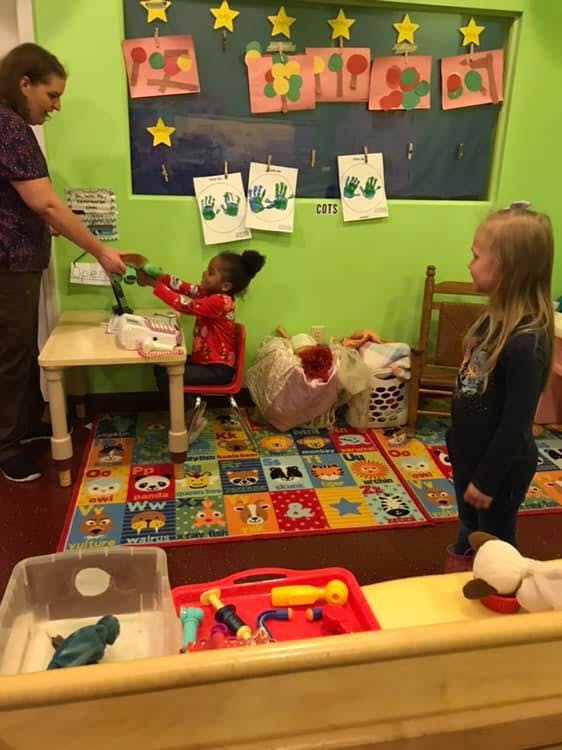 Adult handing an object to a child sitting at a table in a classroom