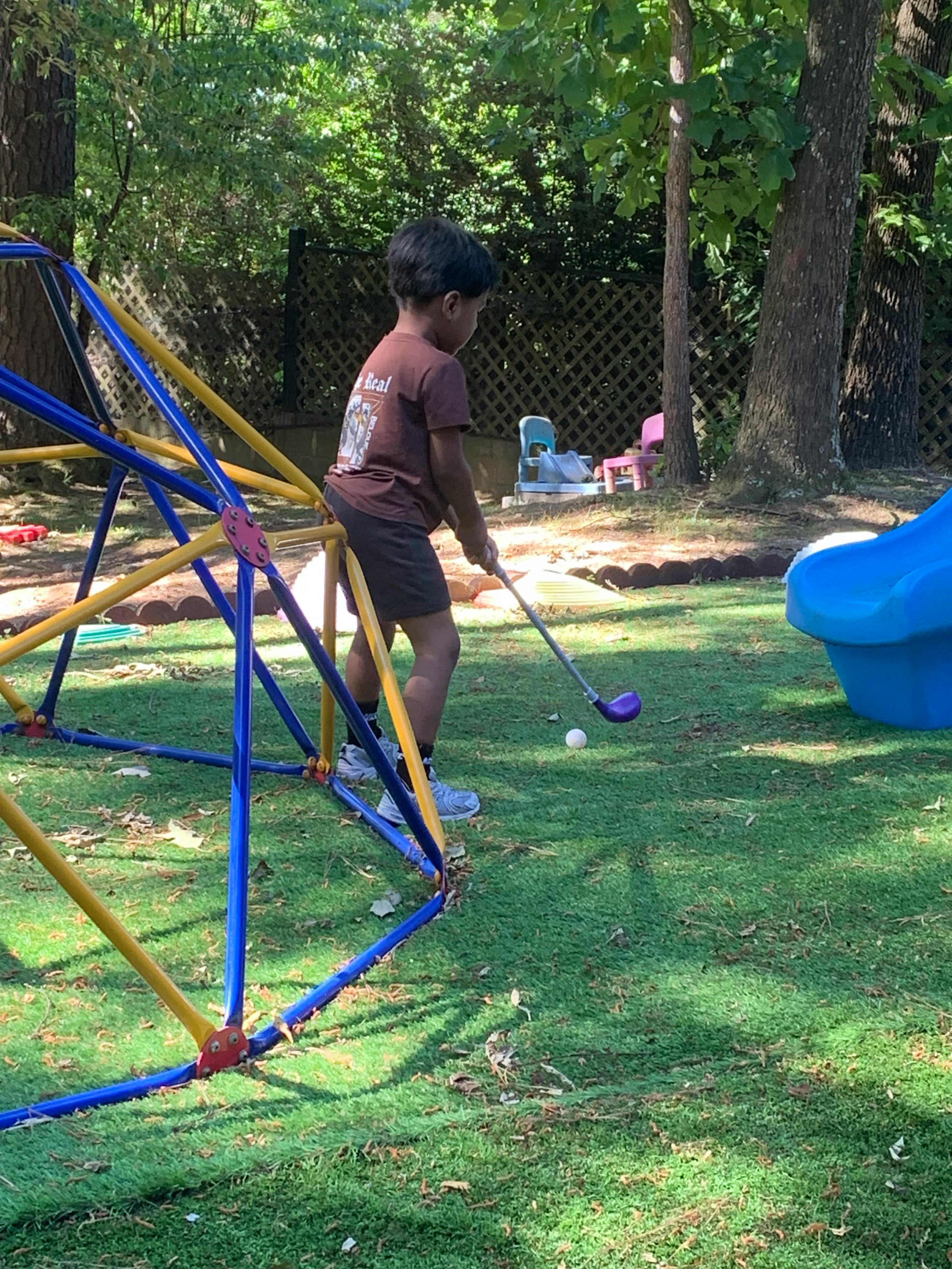 Child playing golf on a jungle gym with a purple golf club and white ball