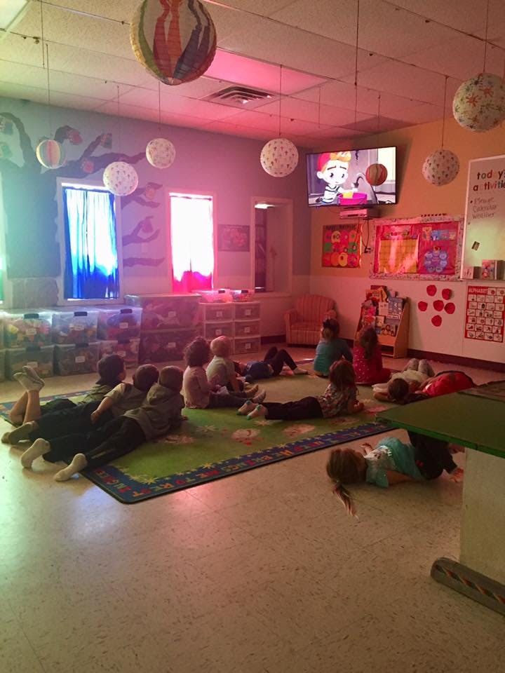 Children sitting and lying on a rug watching a television in a classroom