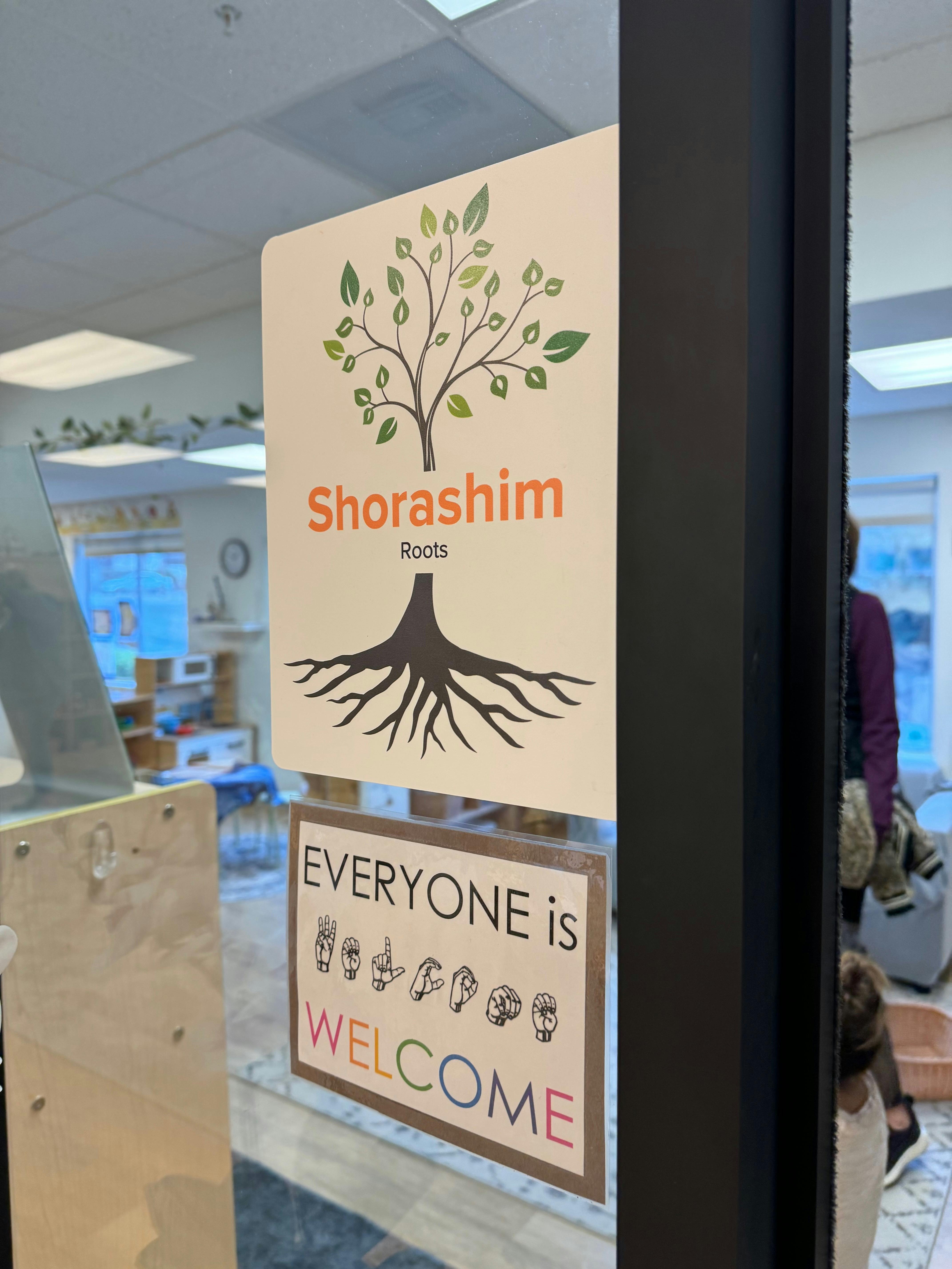Sign on a glass door with a tree logo and text "Shorashim Roots" and "Everyone is Welcome"