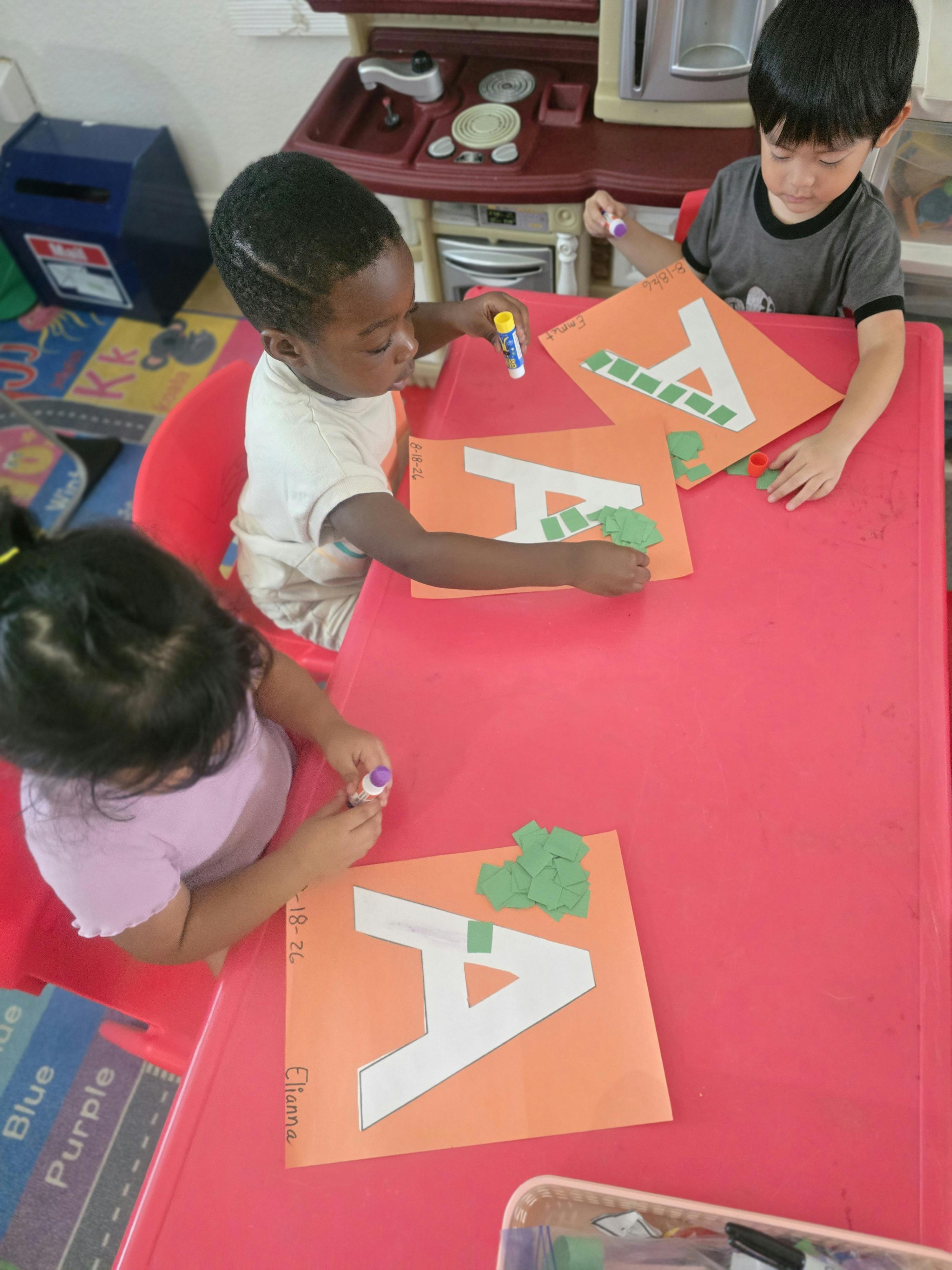 Three children crafting letters on paper at a red table