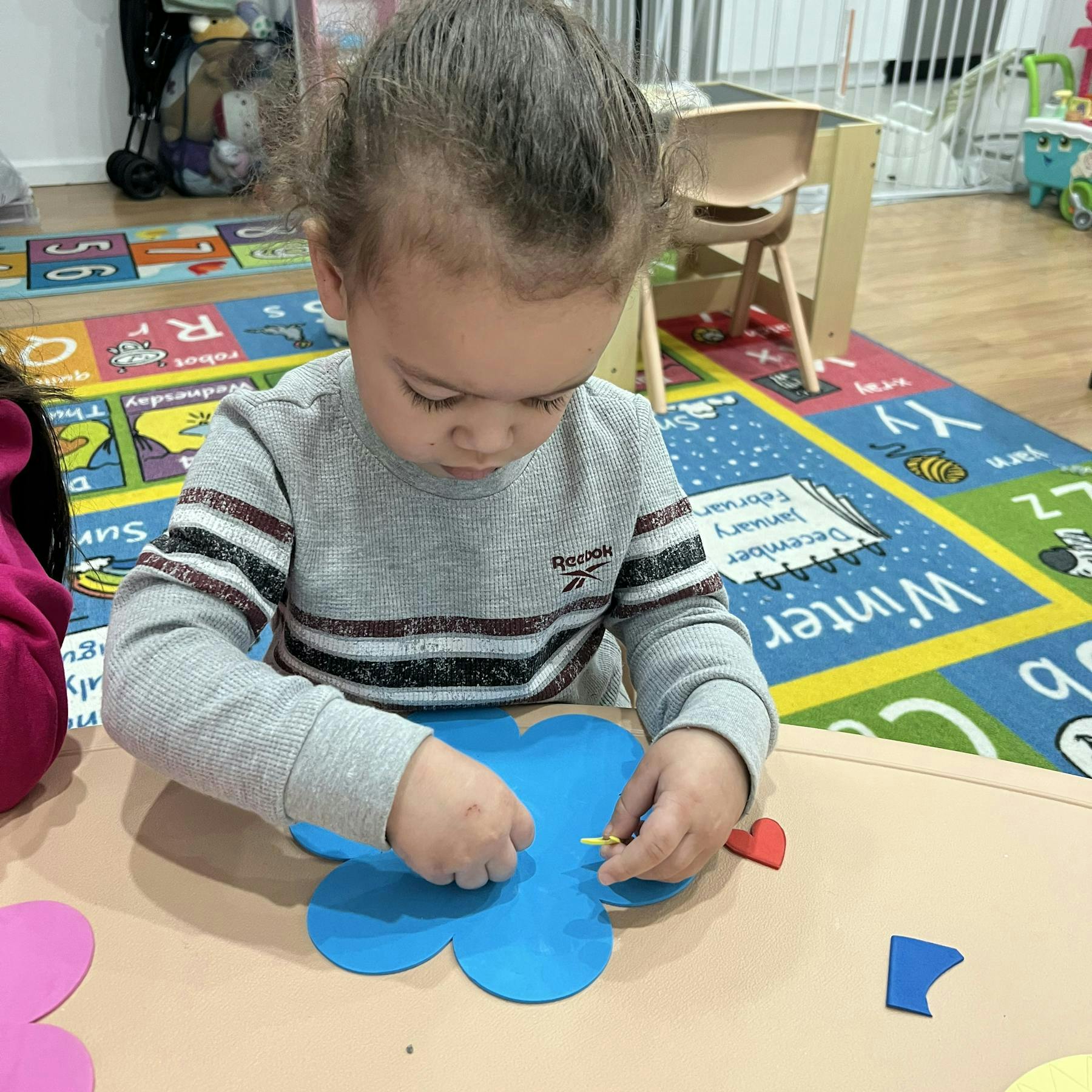 Child manipulating a blue craft shape on a table in a classroom