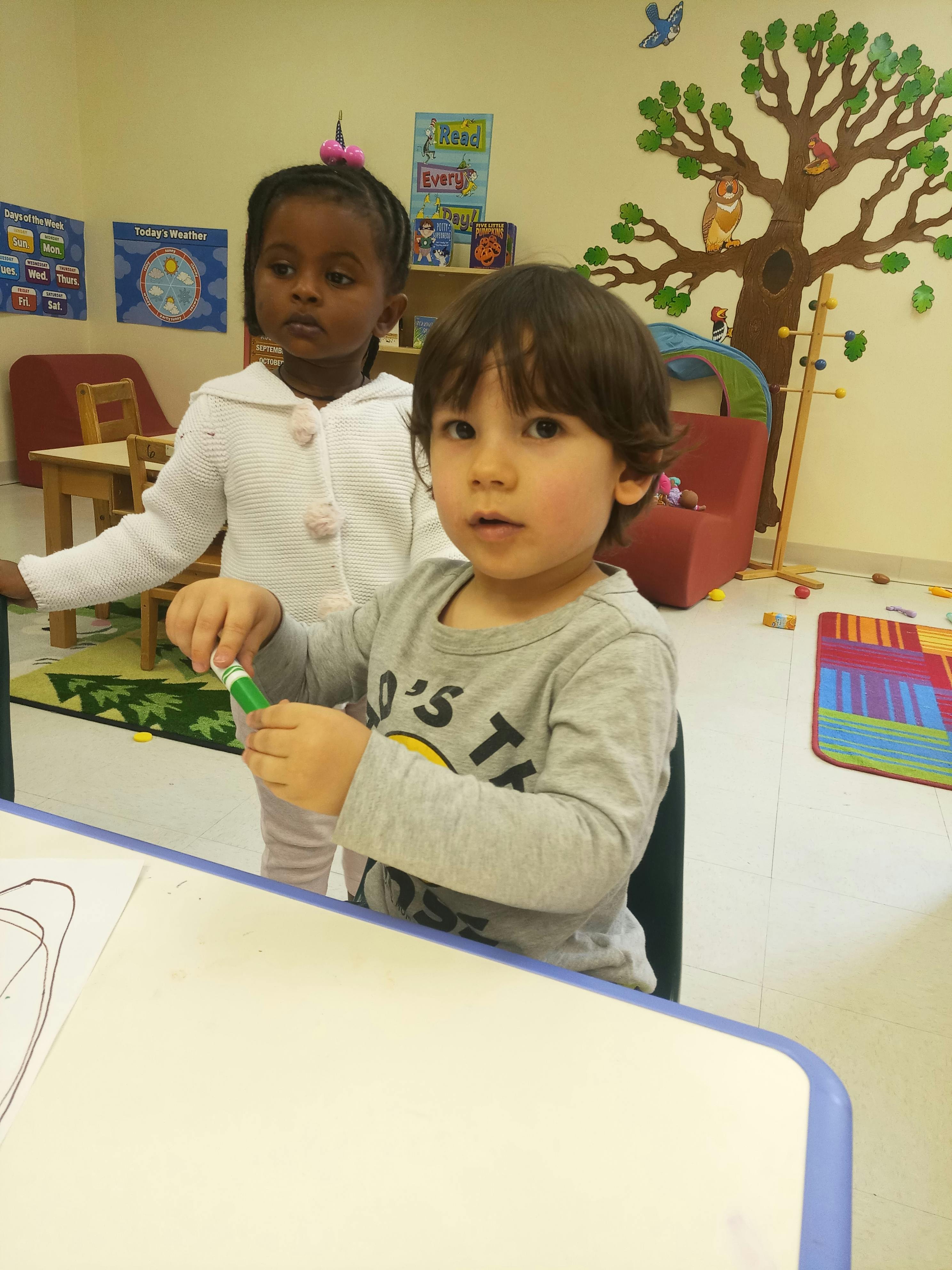 Child holding a marker at a table with another child standing behind