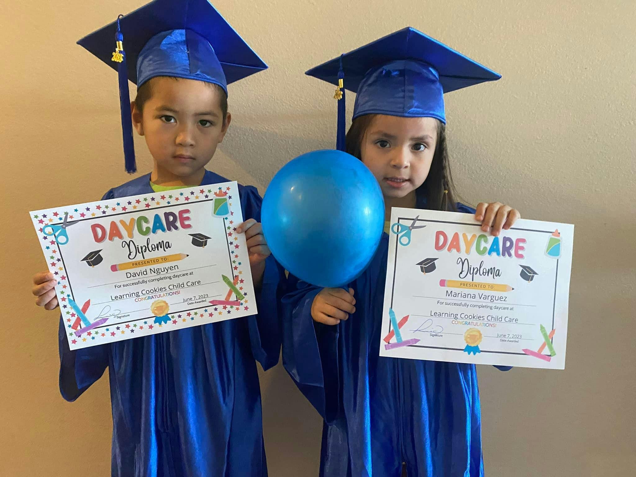 Two children in graduation caps and gowns holding diplomas and a balloon